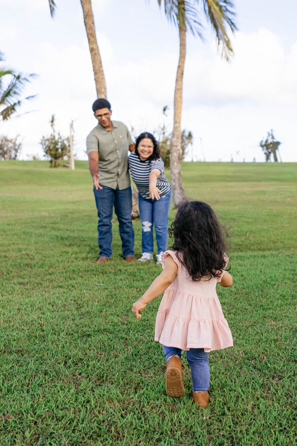 Young girl in pink dress runs toward smiling parents on lush green lawn with palm trees in background.