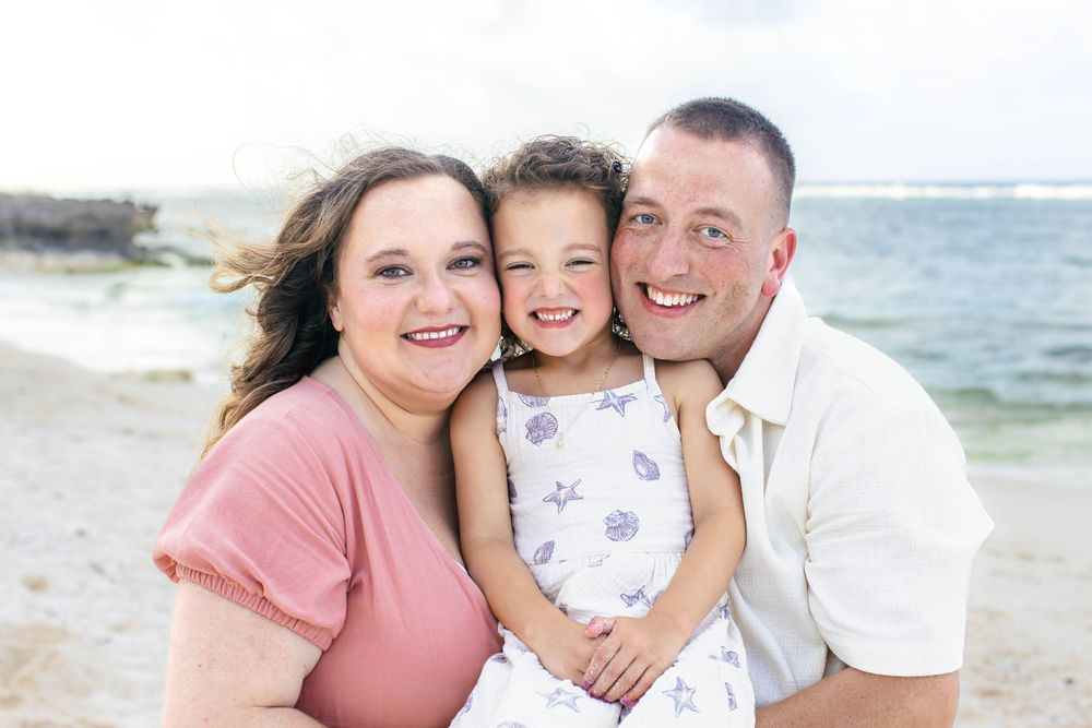 Happy family of three smiling together on a sunny beach, parents hugging young daughter in floral dress.