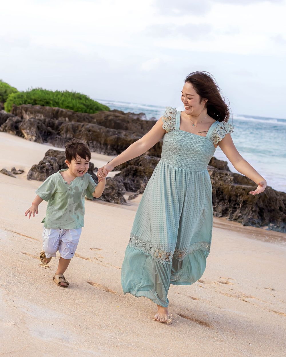 Mother and child running together on a sandy beach with rocky formations and ocean waves in the background.