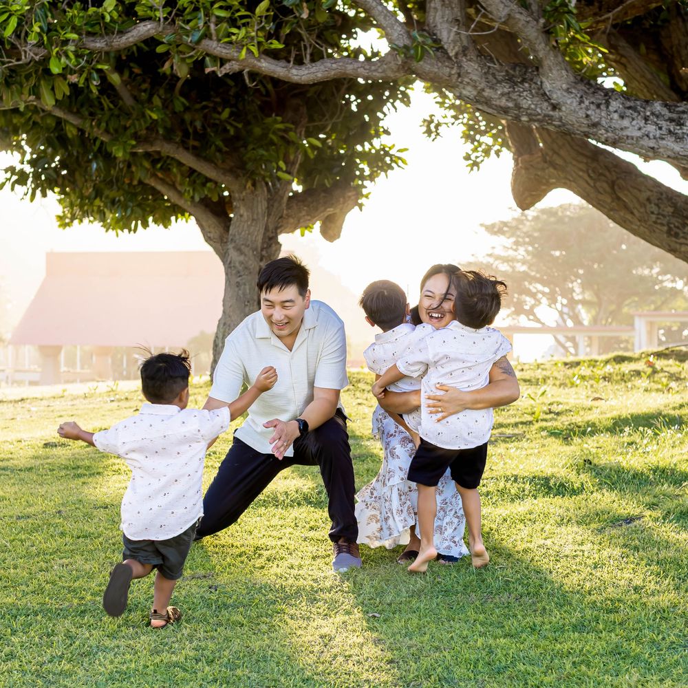 Family playing outdoors under a large tree on green grass during golden hour in a tropical park setting.