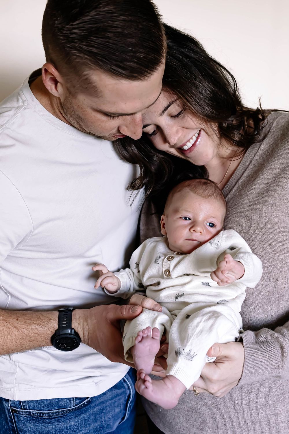 Happy couple smiling and holding their newborn baby dressed in white onesie, sharing a tender family moment.