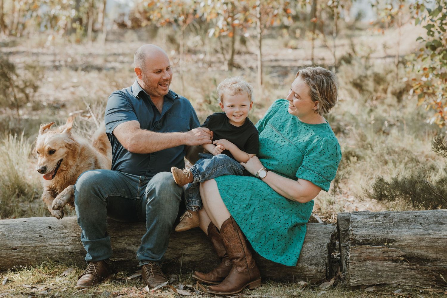 Family with young child and golden retriever sitting on log in autumn vineyard setting.