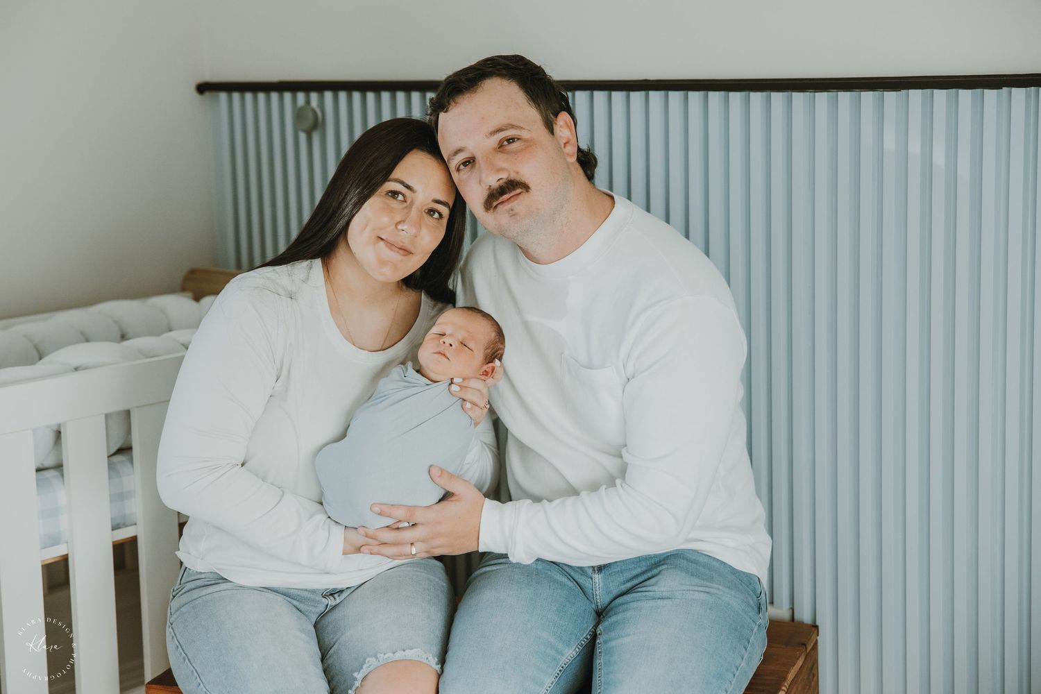 Happy couple in white sweaters and jeans holding their newborn baby in a light blue nursery room.
