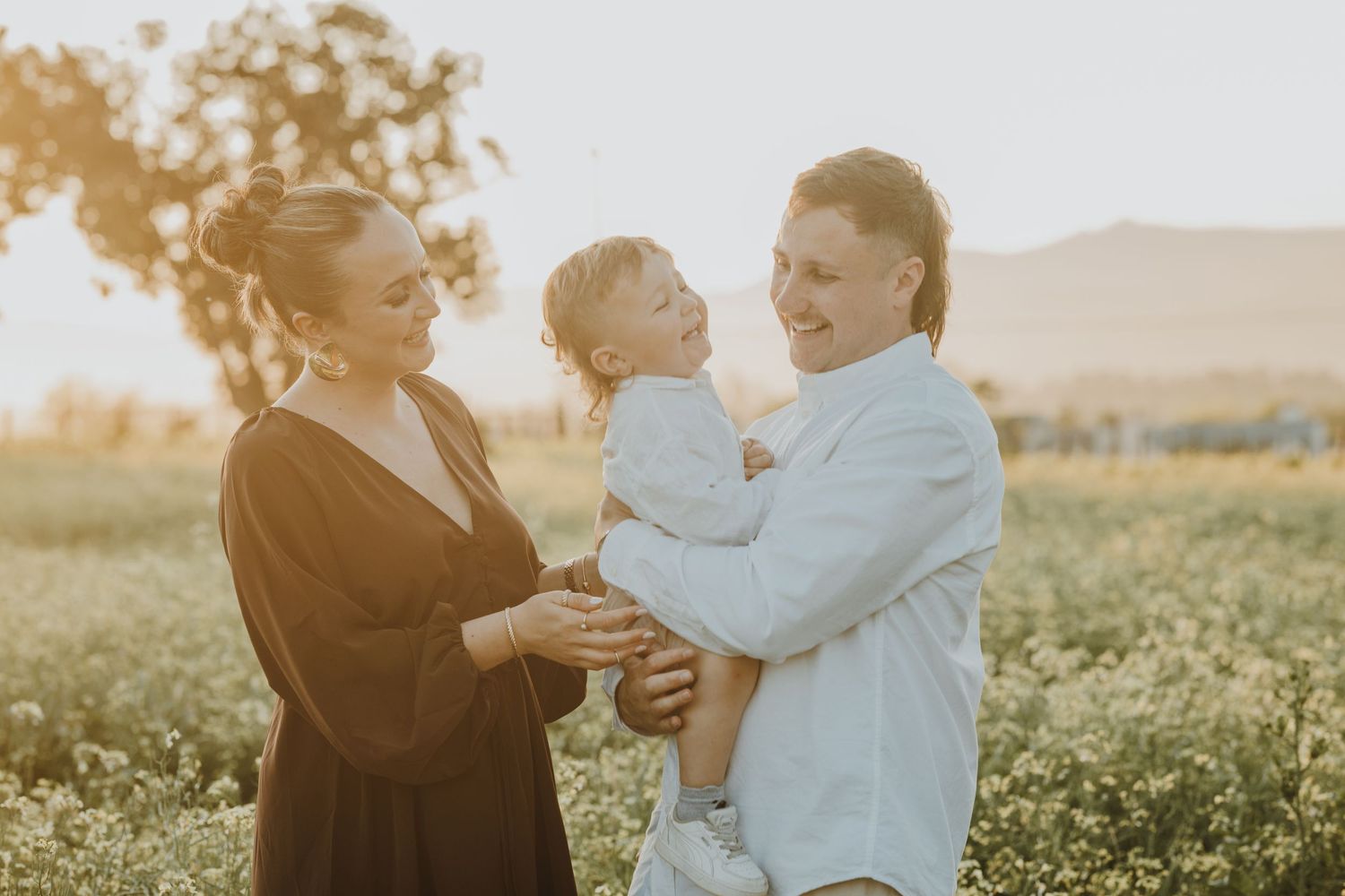 Family of three enjoying golden hour in a blooming field, parents smiling at toddler in white outfit.