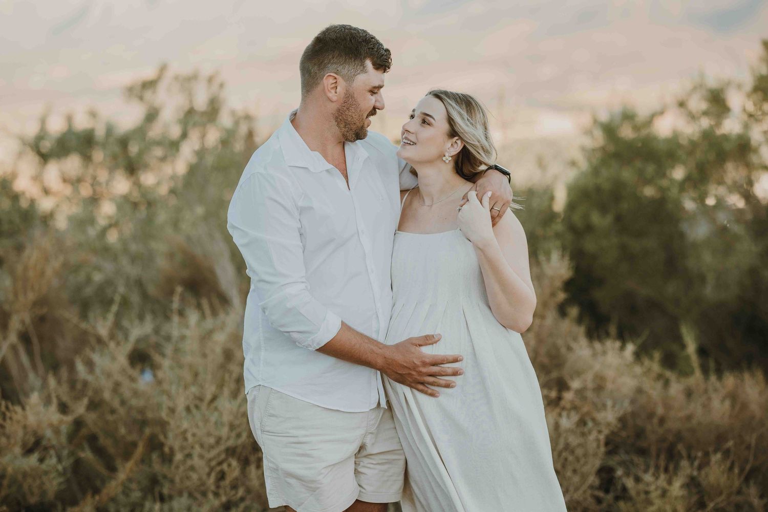 Couple in matching white outfits embracing outdoors with mountains and greenery in the background during golden hour.
