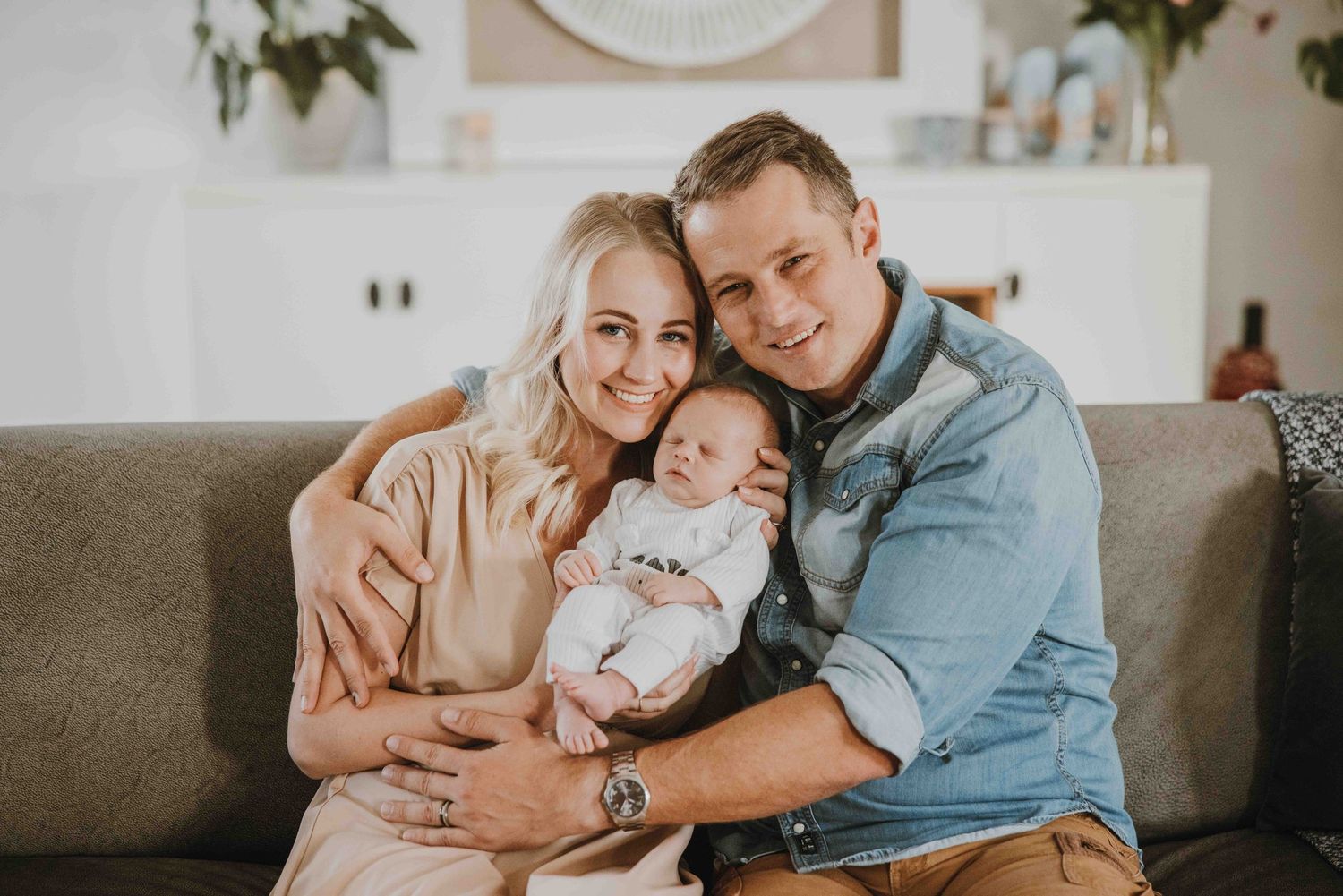 Happy young couple sitting on a couch smiling while holding their newborn baby in a bright living room.