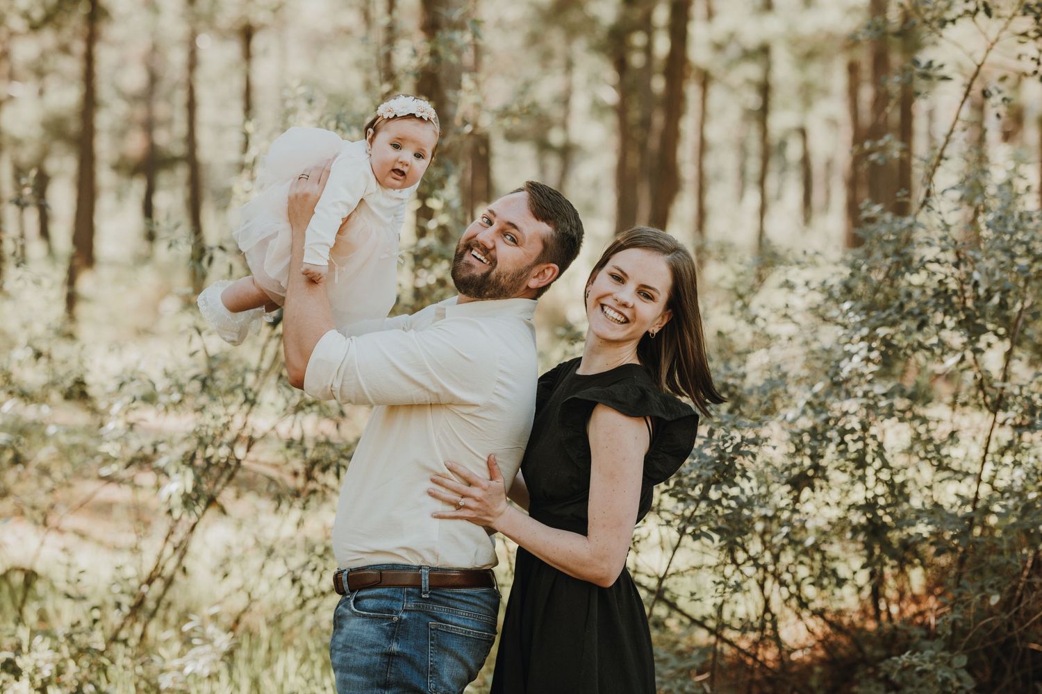 Happy young family with baby in a sunlit forest, parents smiling and holding infant during outdoor photo session.
