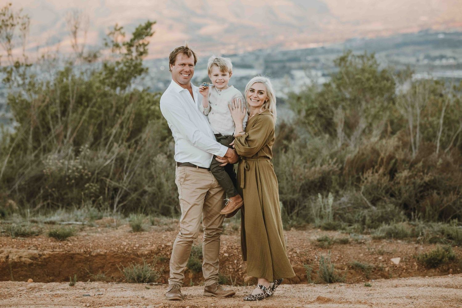 A happy family of three poses outdoors at sunset, with parents holding a toddler against a scenic mountain backdrop.