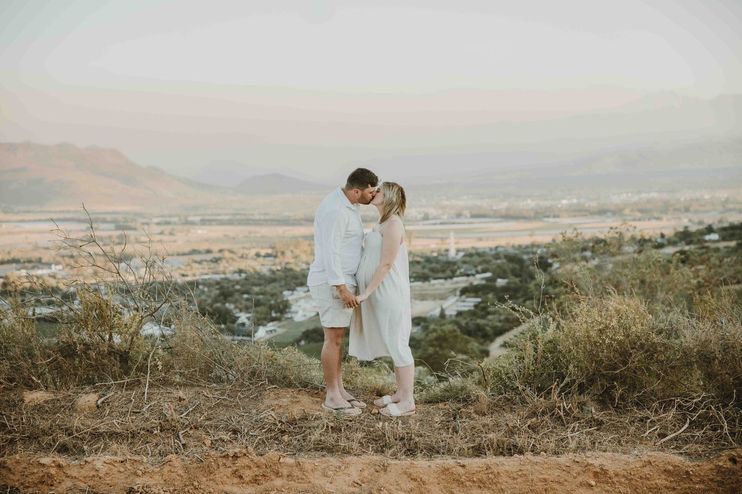 Couple sharing a romantic kiss on a scenic hilltop with mountain valley views in soft golden light.