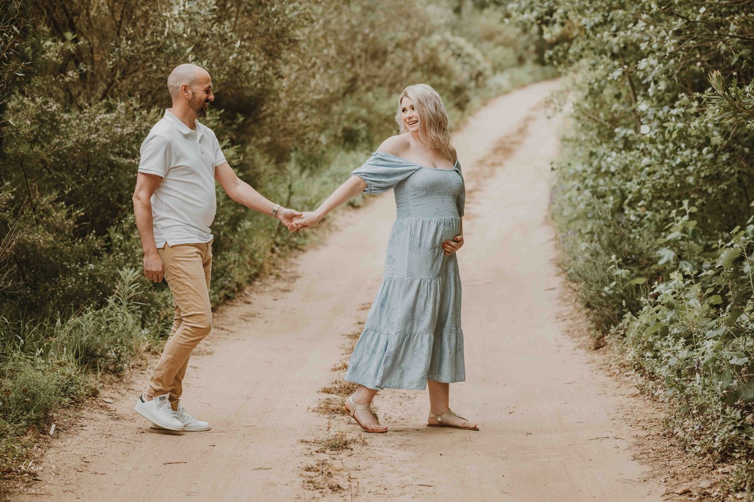 Pregnant couple holding hands on a scenic dirt path surrounded by lush greenery, wearing casual summer outfits.