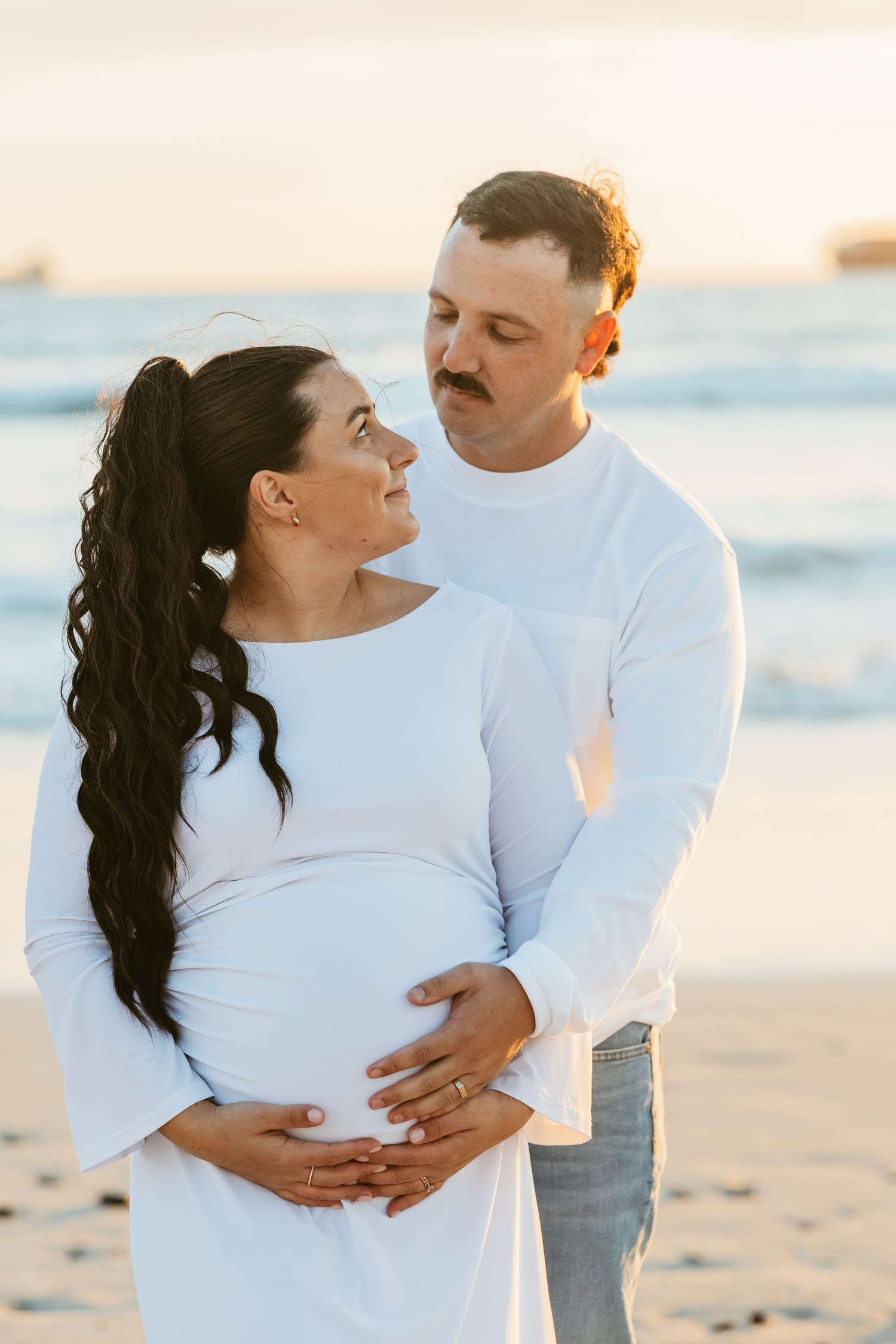 Couple in white outfits on beach at sunset, man embracing pregnant woman from behind, both smiling tenderly.