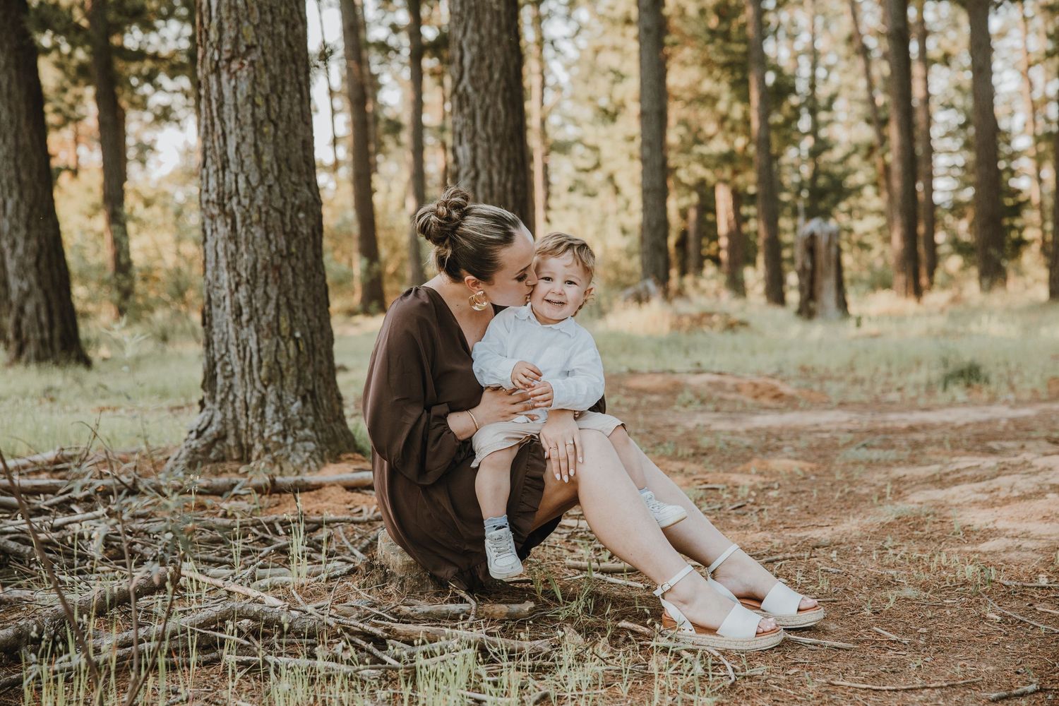 A mother kisses her toddler while sitting together in a pine forest, sharing a tender outdoor moment.