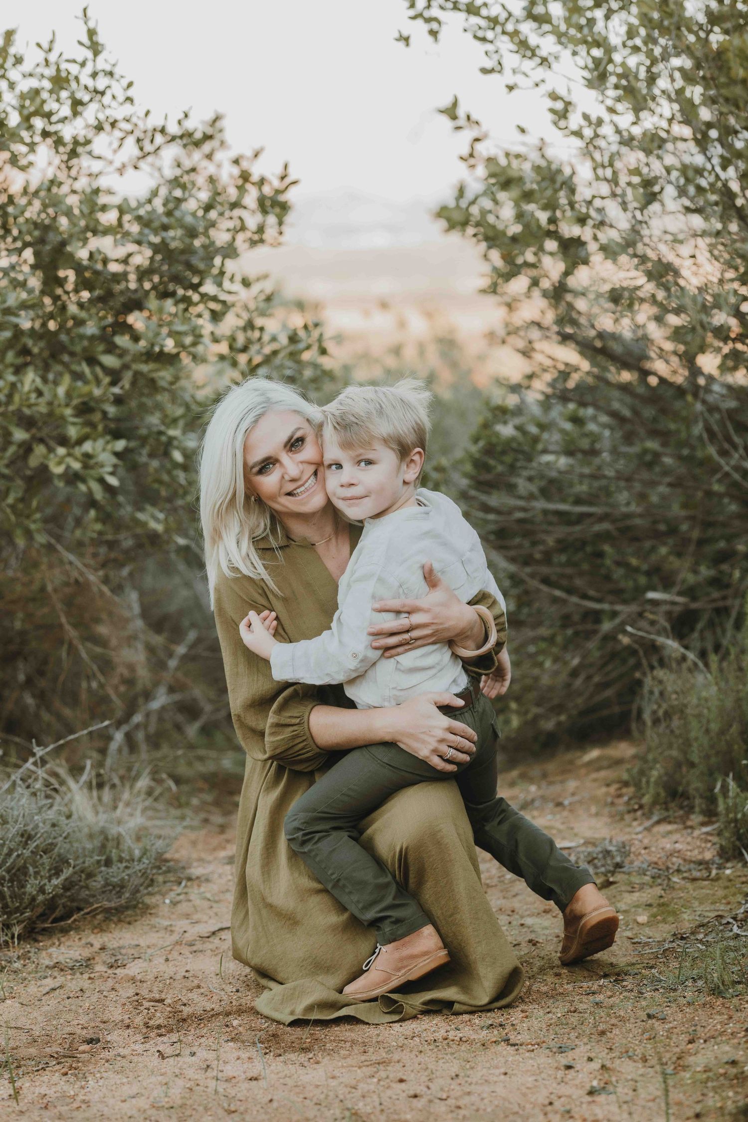 A smiling blonde woman in an olive dress holds a young child outdoors among green shrubs on a dirt path.