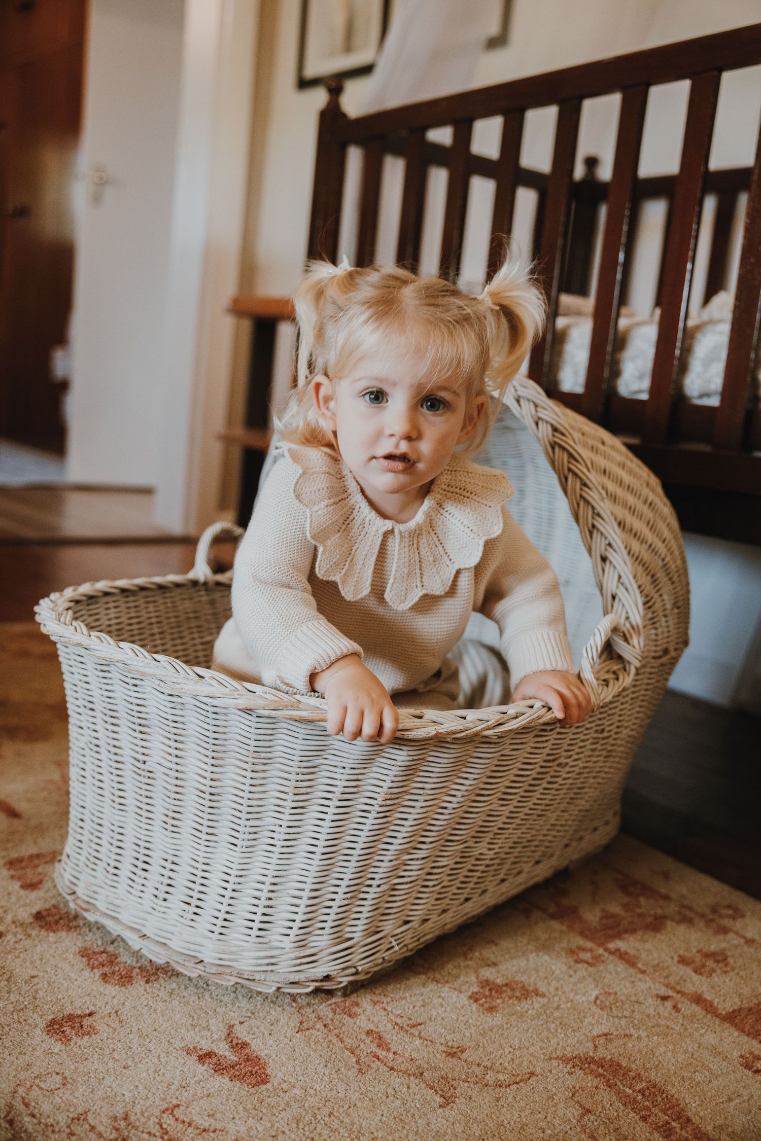 A toddler in a cream dress sitting inside a white wicker basket on a patterned rug.
