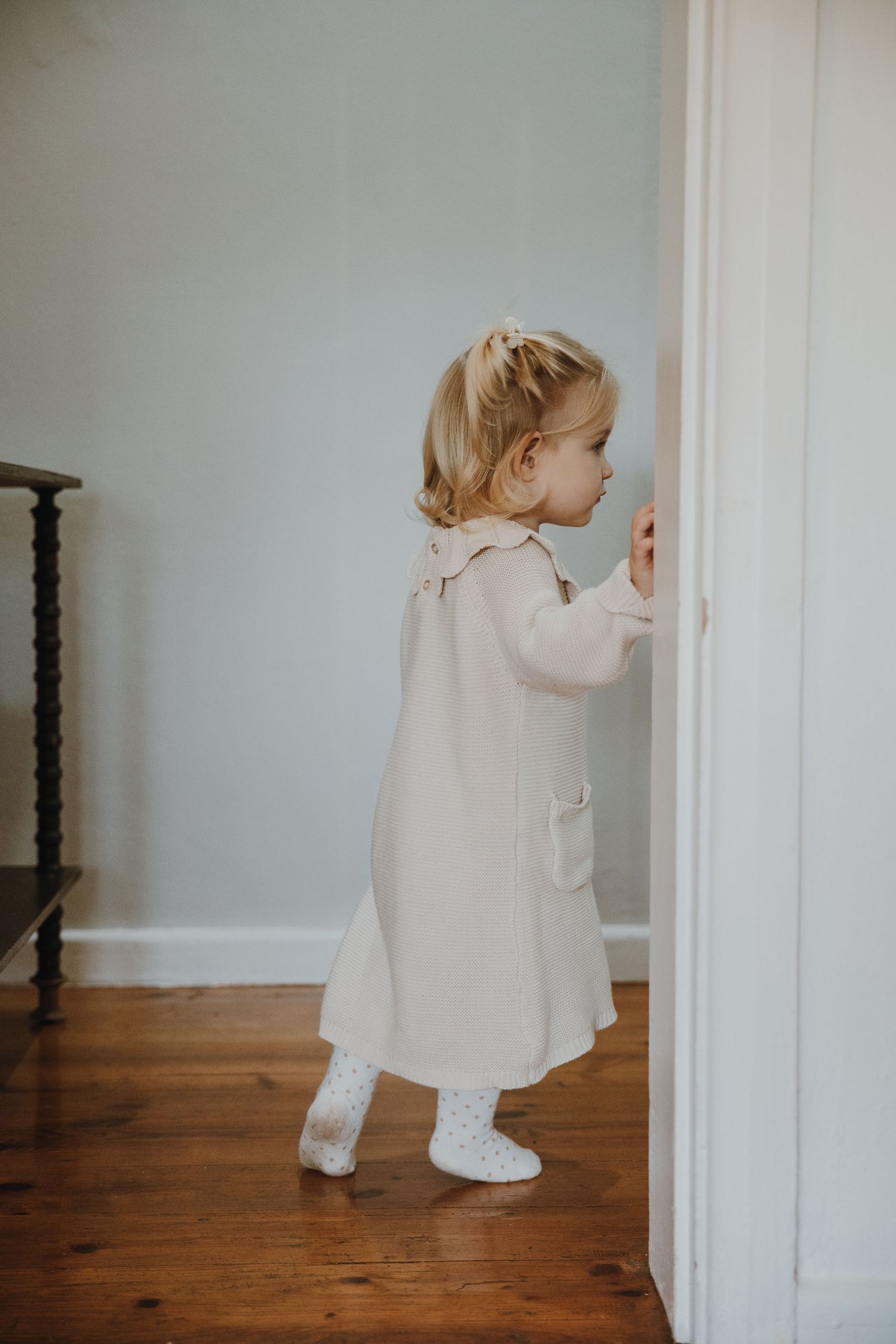 A toddler in a cream-colored dress reaching for a white door handle on hardwood floors.