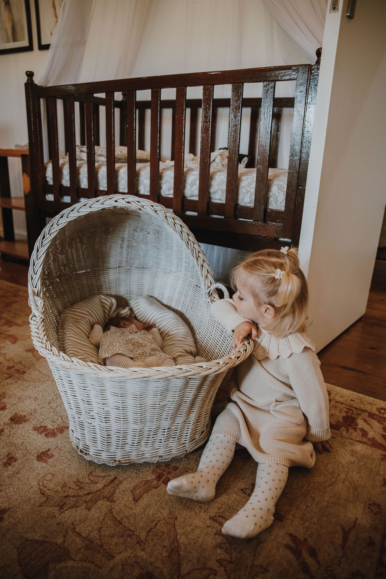 A view of a nursery with a dark wood crib and white wicker basket on a patterned rug.
