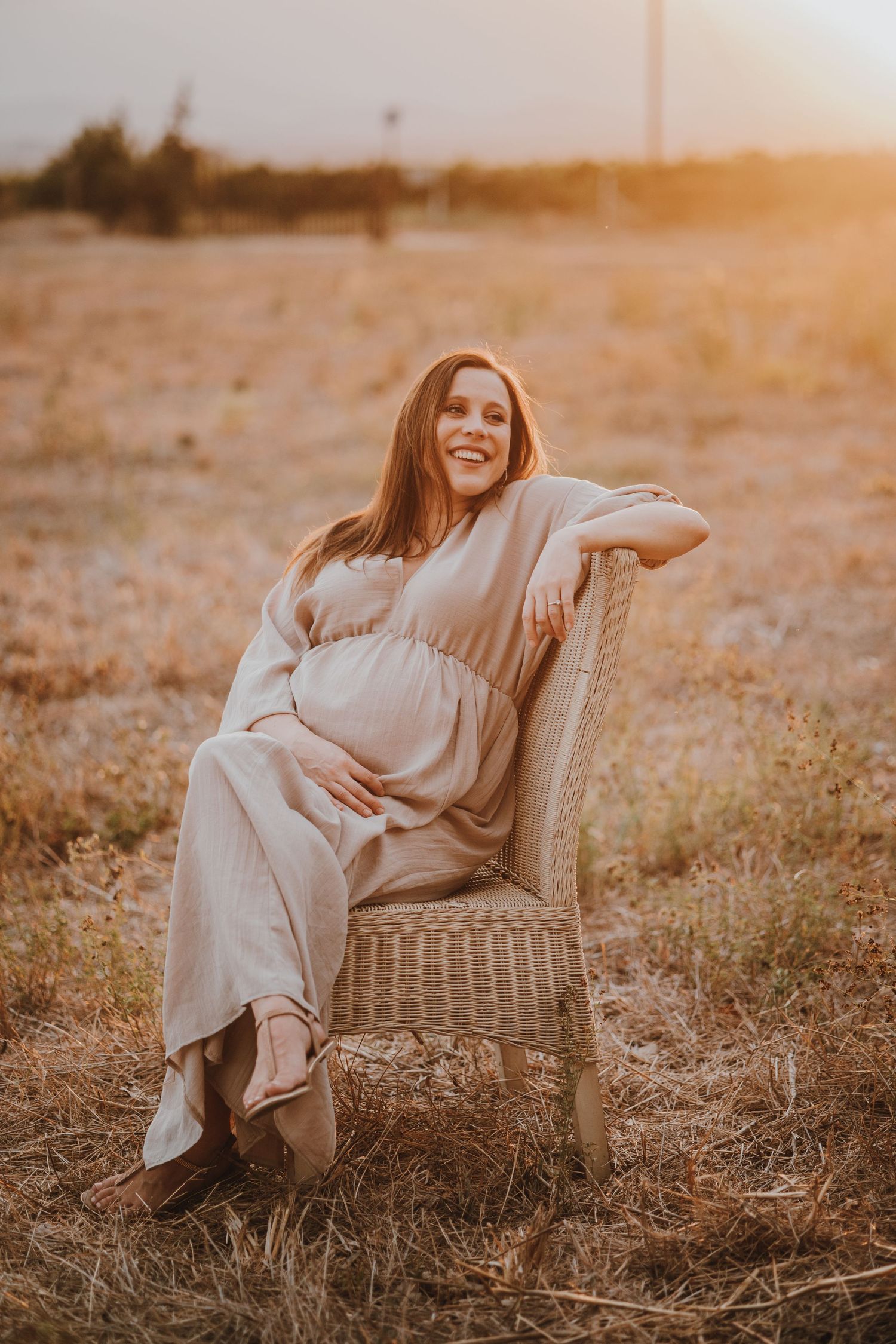 Expectant mother in a flowing white dress sits in wicker chair during golden hour sunset in field.