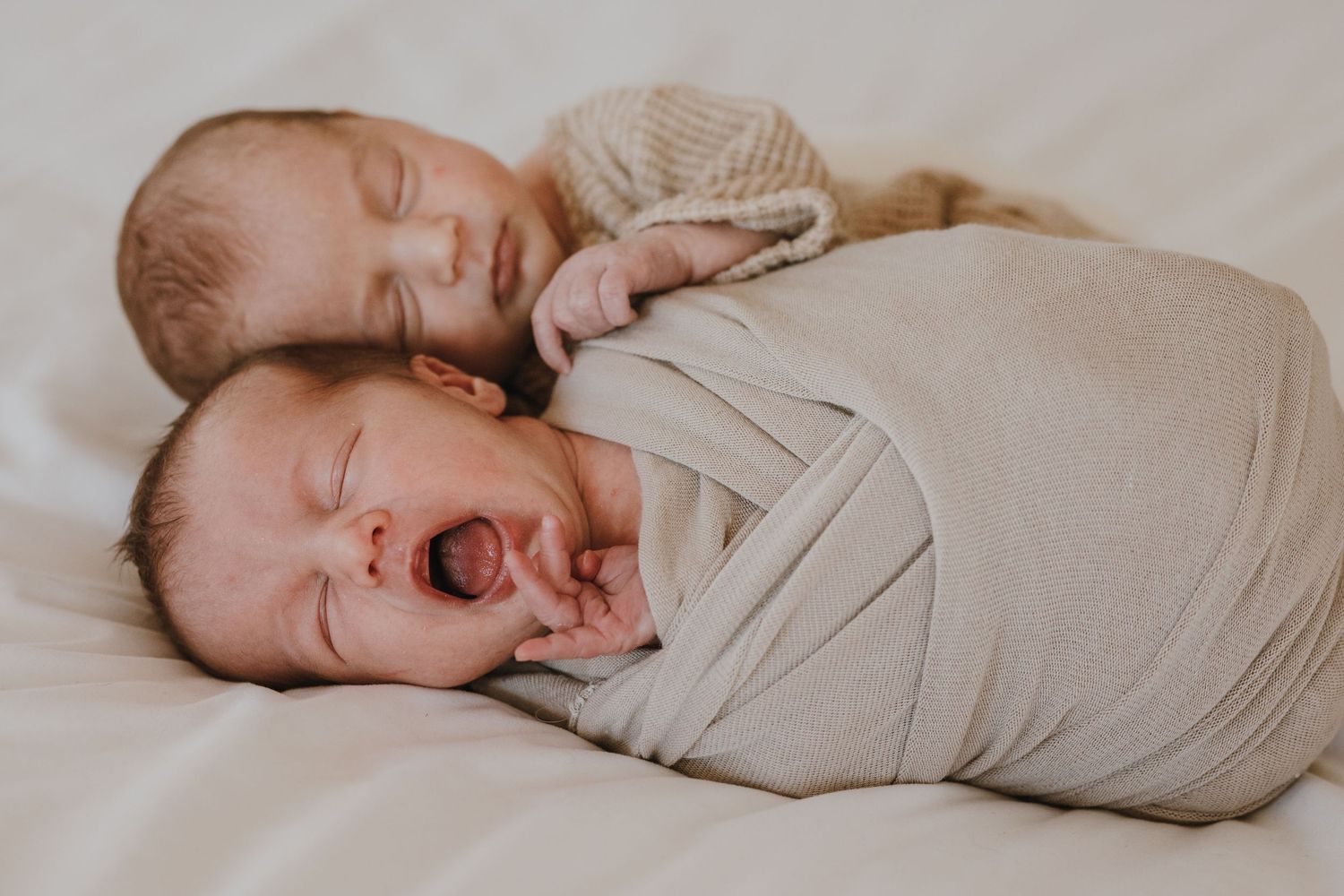 Two wrapped babies on white bedding, one sleeping and one crying.