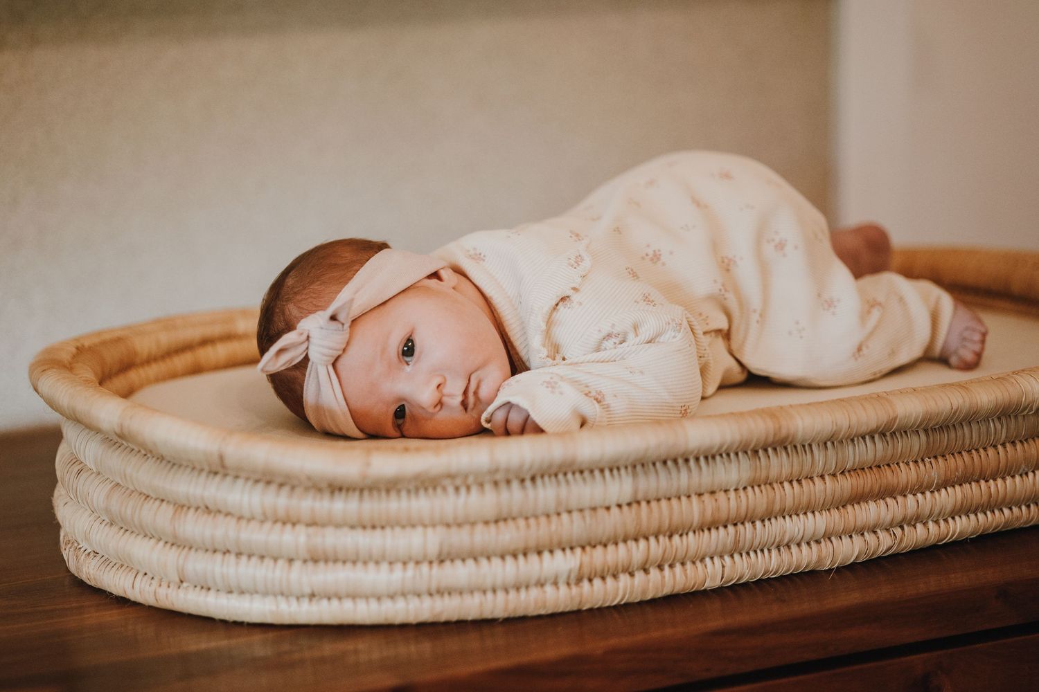 A baby in white clothing lying in a woven basket on a wooden surface.