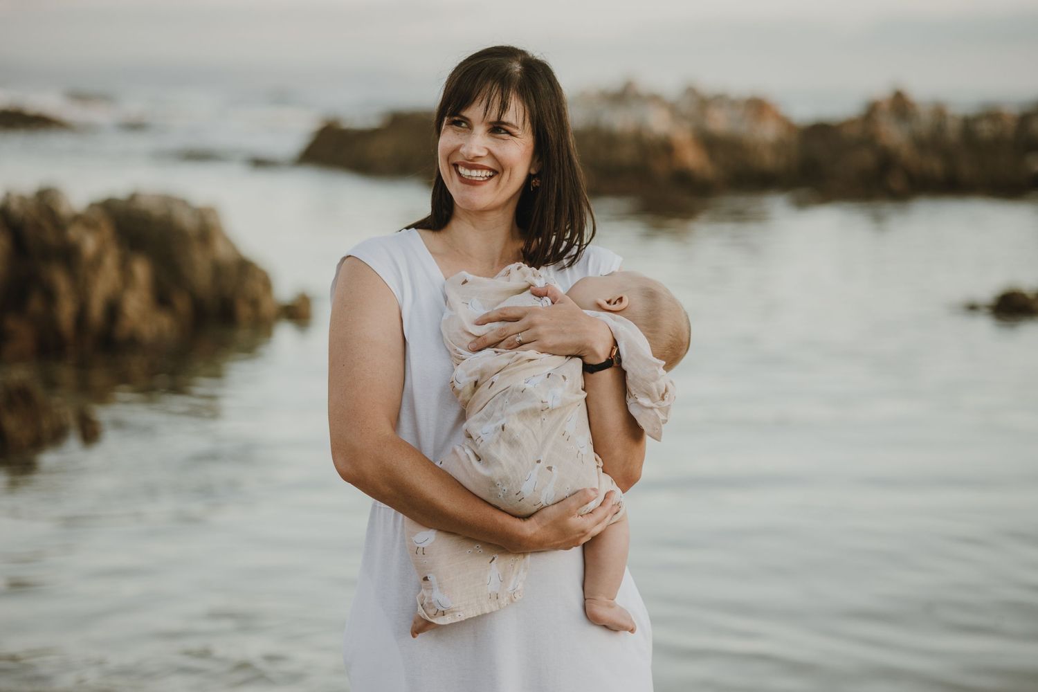A tender moment between parent and infant at a coastal setting with rocky outcrops.