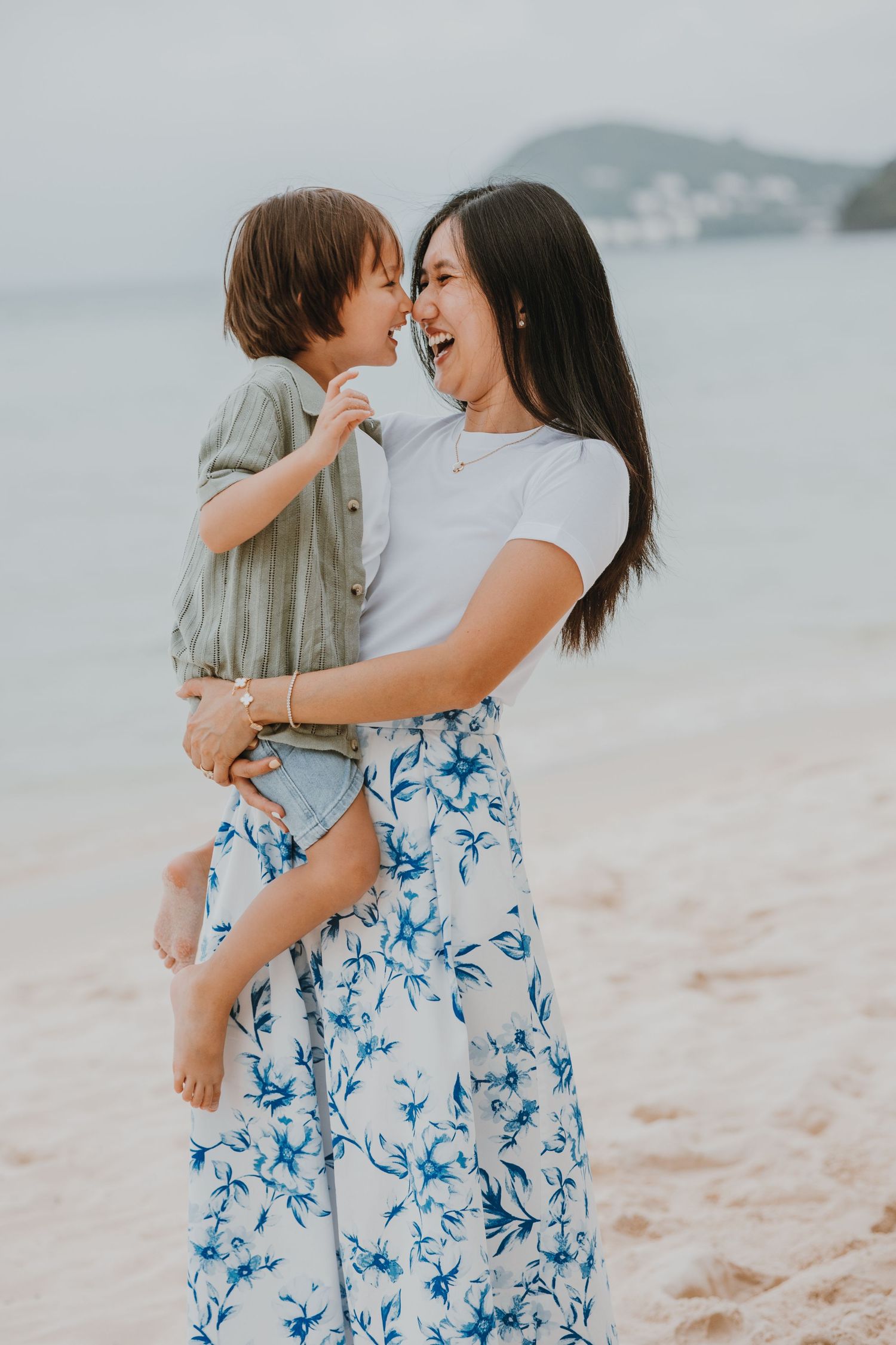 A joyful moment shared between parent and child on a misty beach, wearing a flowing blue floral skirt and white t-shirt.