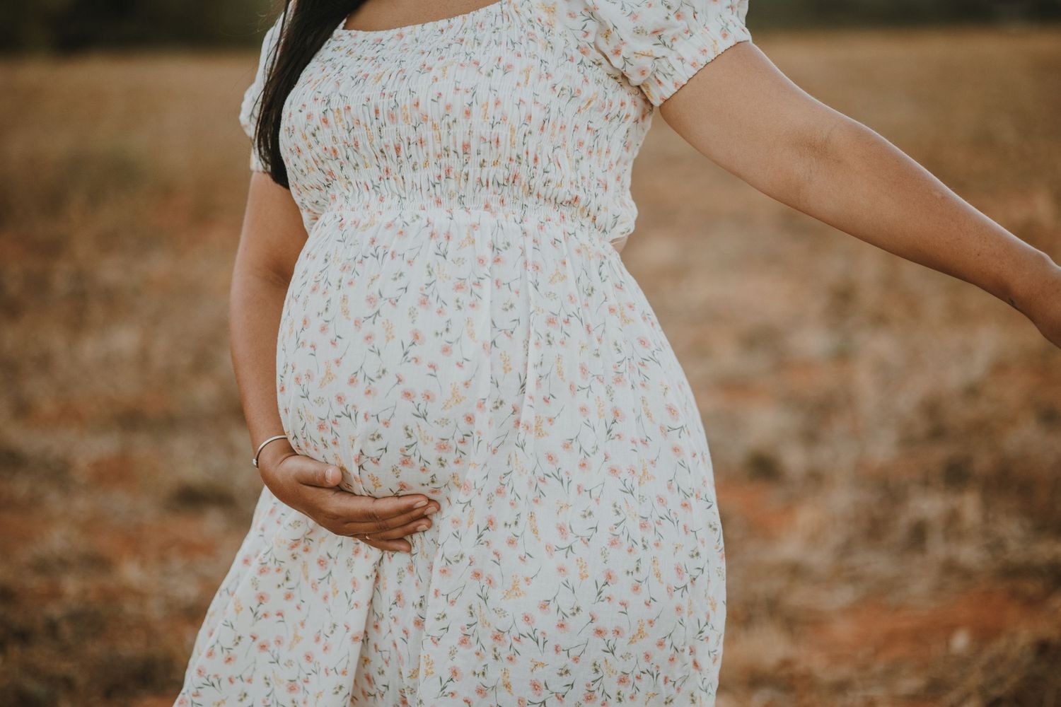 A pregnant woman in a floral white dress stands in a sunlit field during golden hour.
