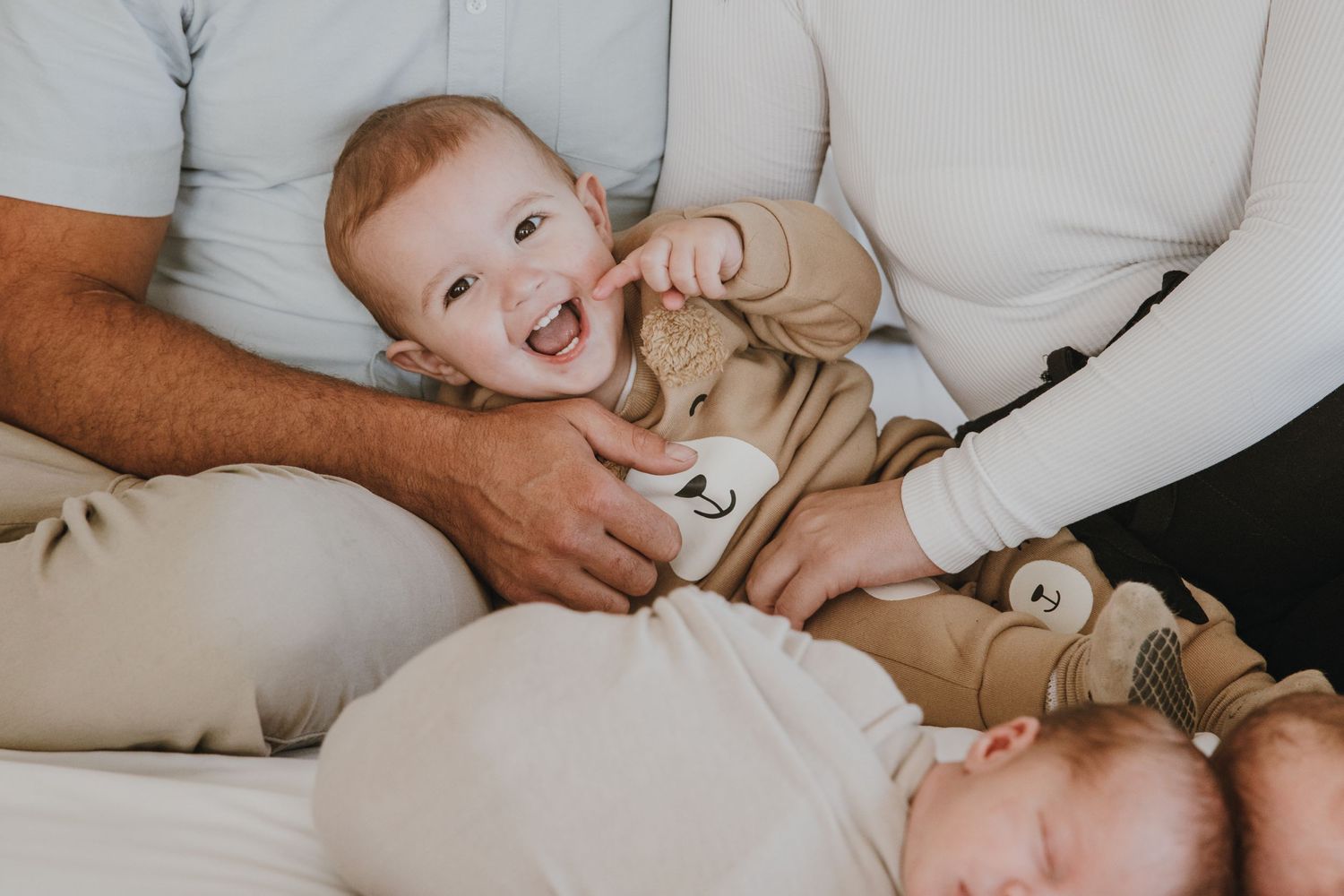 A laughing baby in beige clothing plays on white bedding.