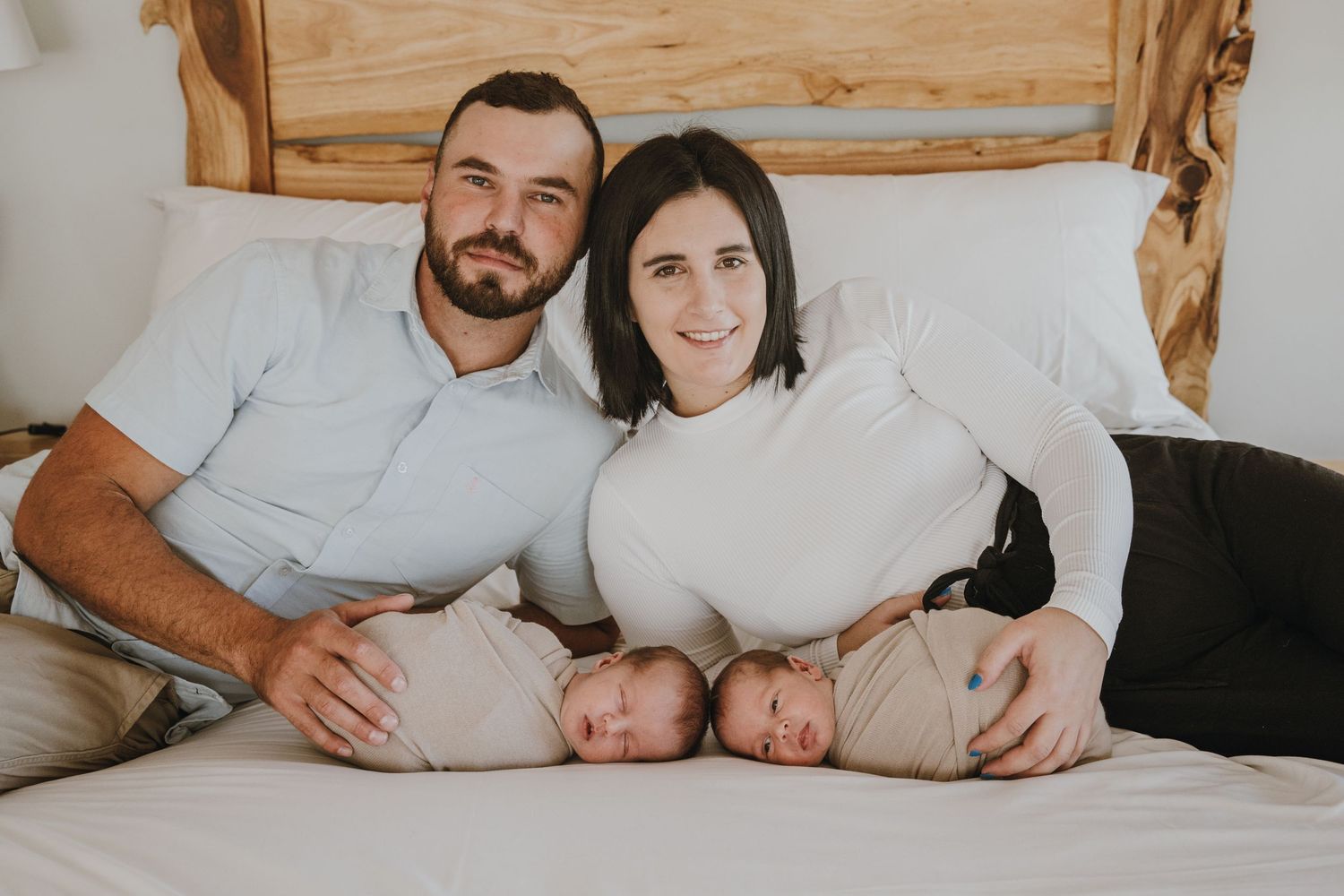 A family with twin babies rests together on a white bed with wooden headboard.