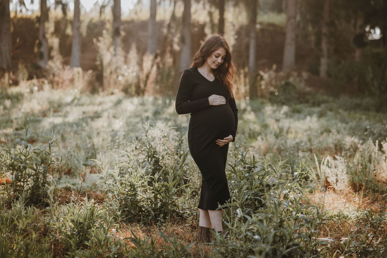 A maternity portrait in a wooded setting with golden sunlight filtering through trees.