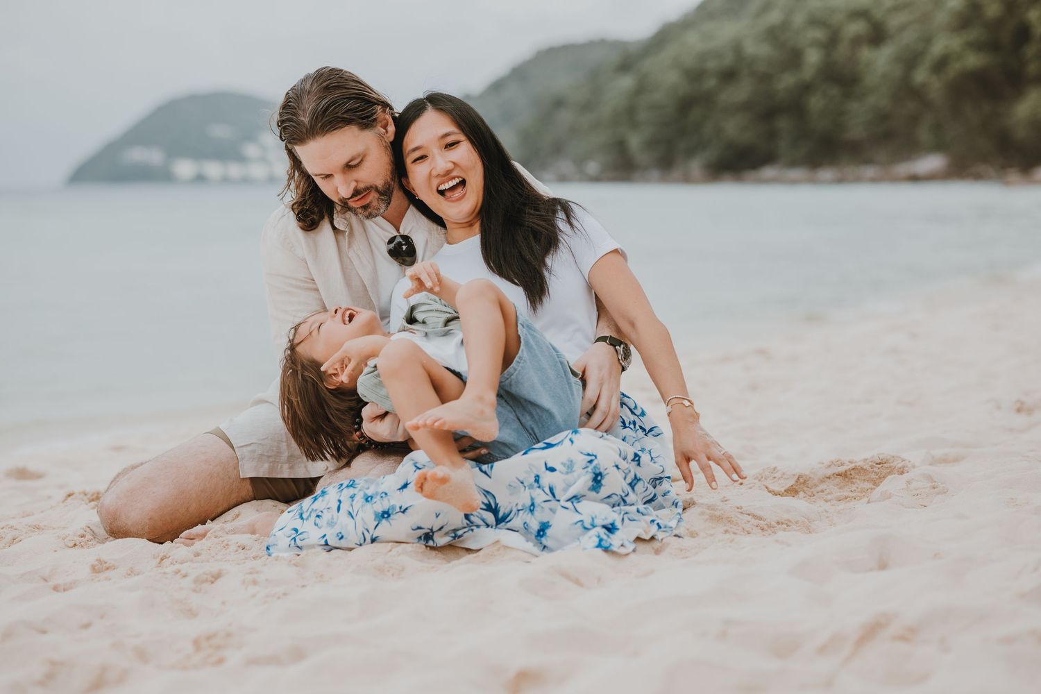 Three people wearing blue and white floral dresses share a joyful moment together on a sandy beach.