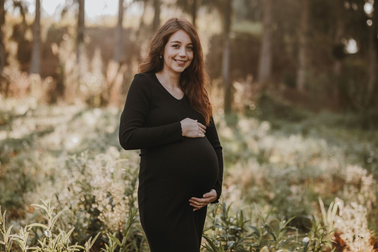 An outdoor maternity photo in a black dress among tall grass and trees at sunset.