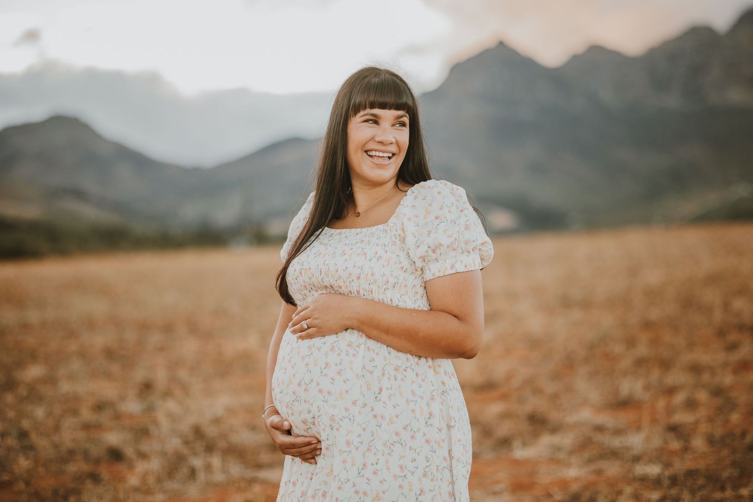 Expectant mother in white lace dress stands in golden field with majestic mountain range in background at sunset.
