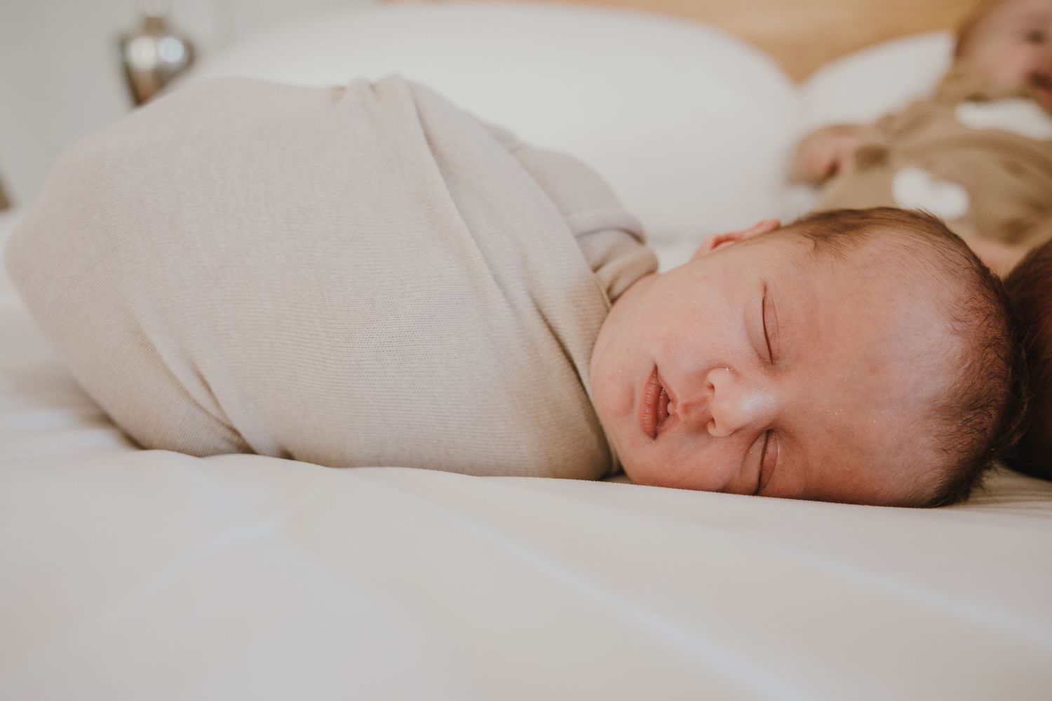 A sleeping infant in beige wrapping rests peacefully on white bedding.