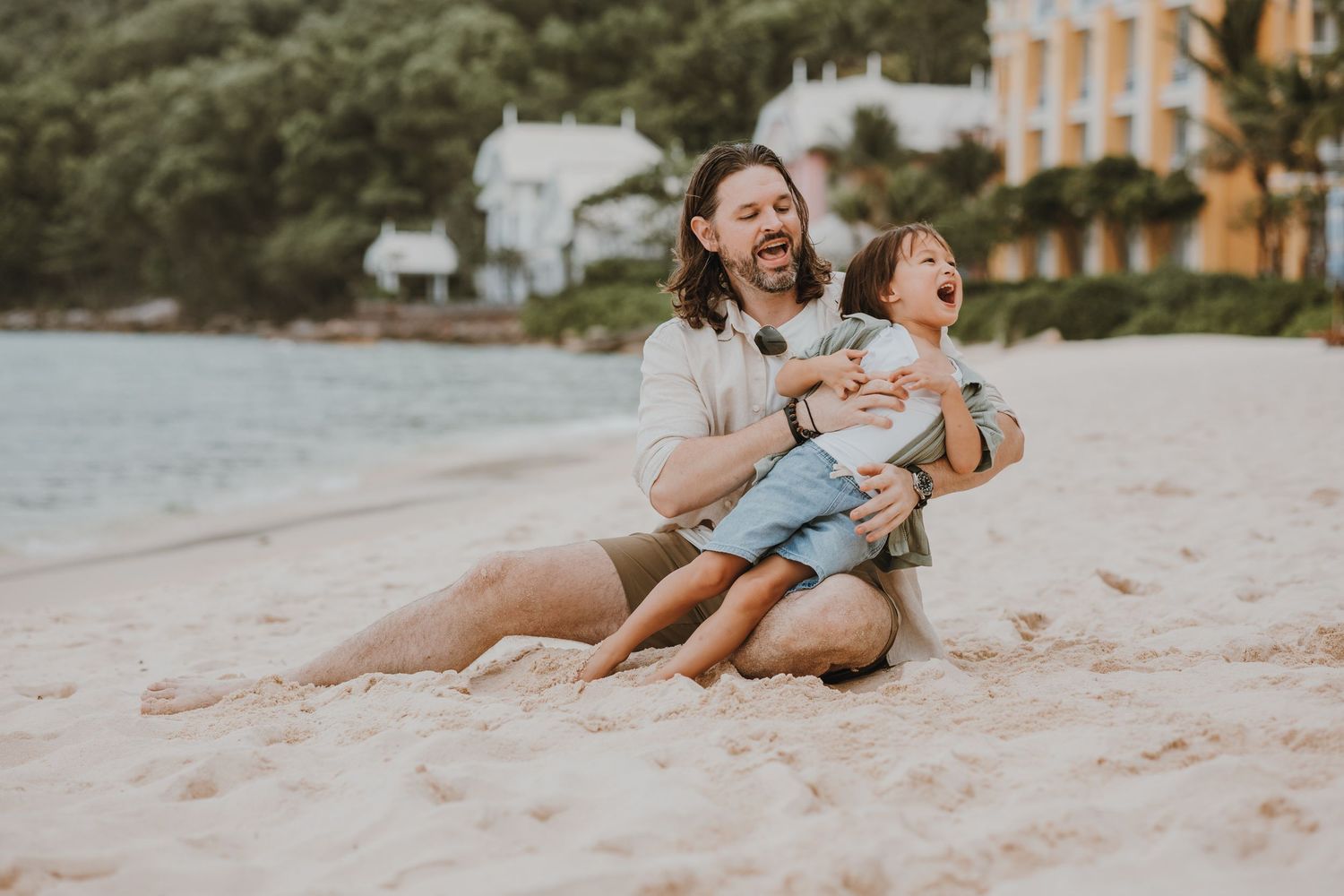 Two people laughing together during a sunset beach photoshoot while sitting on the sand.