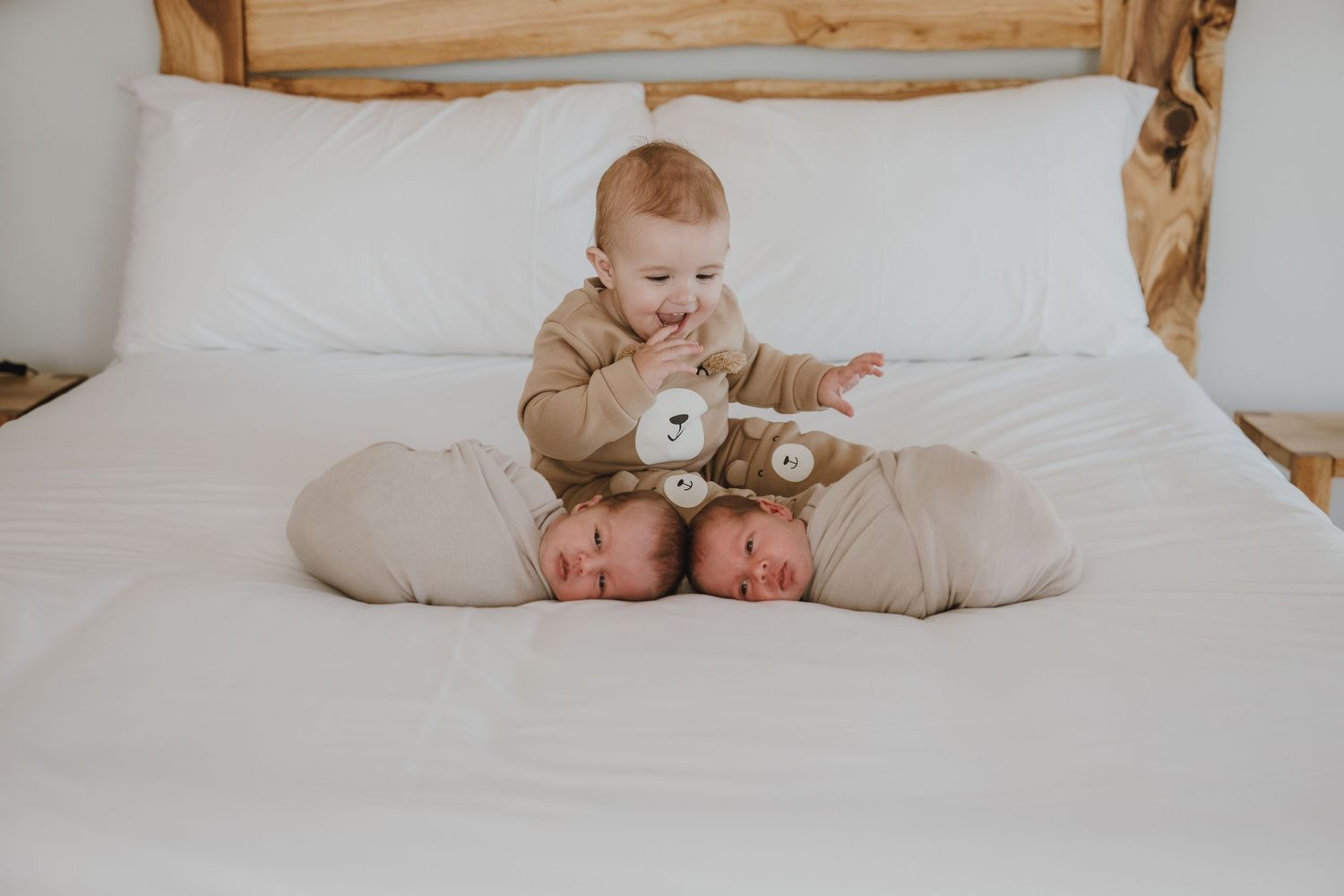 A small infant in a tan onesie sits on white bedding holding a stuffed toy.