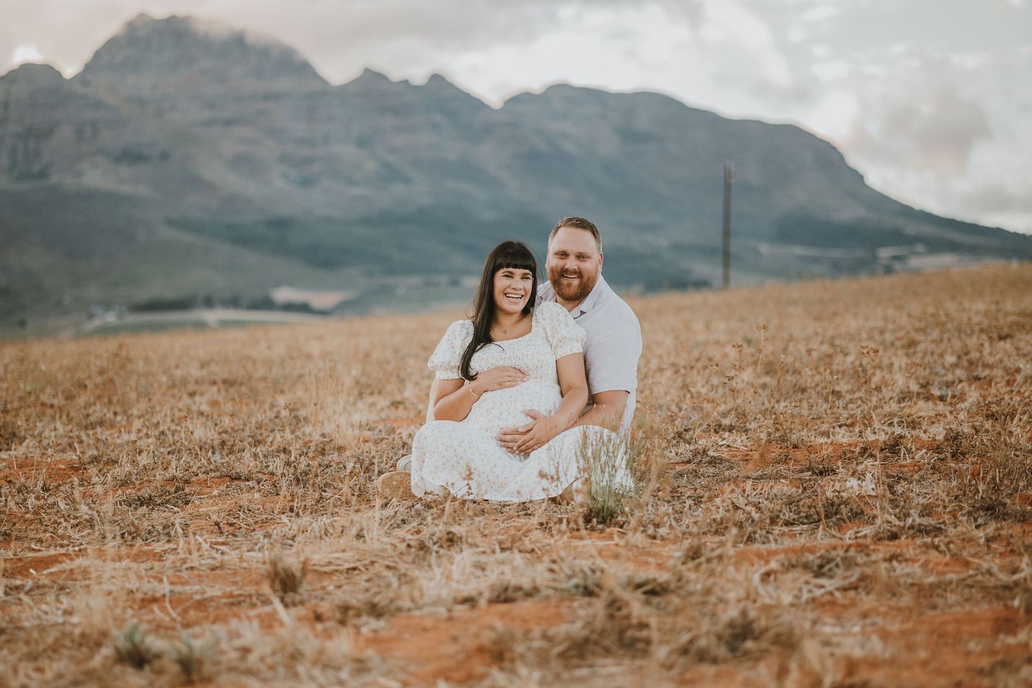 A couple sits in a golden field with dramatic mountain peaks rising in the background during sunset.
