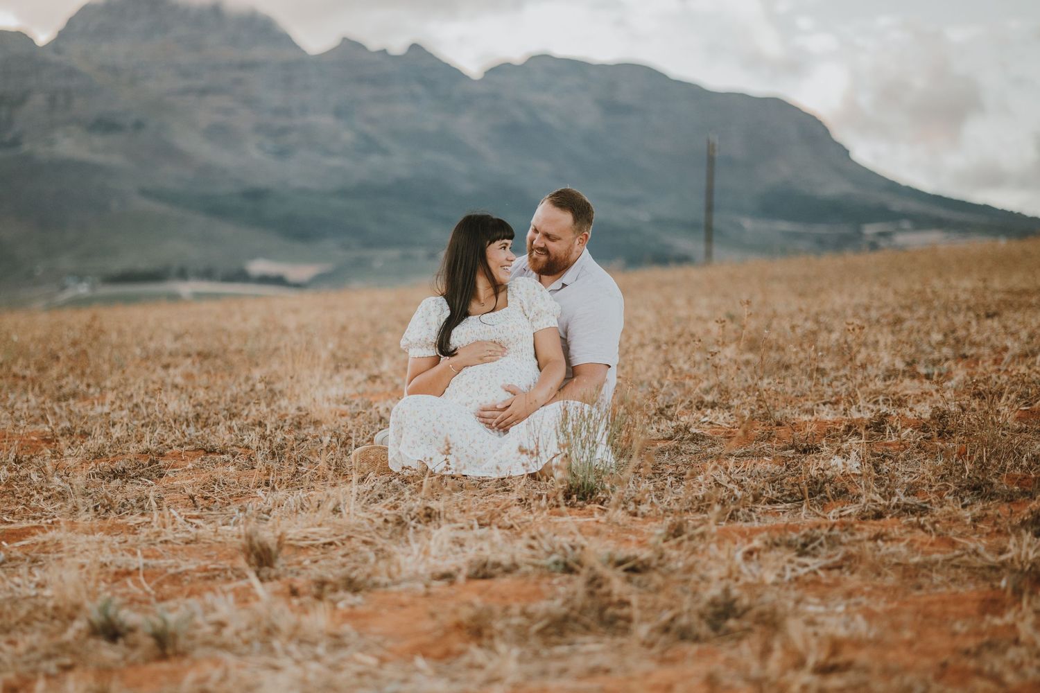 A couple embraces while sitting in a dry grass field against a mountainous backdrop.