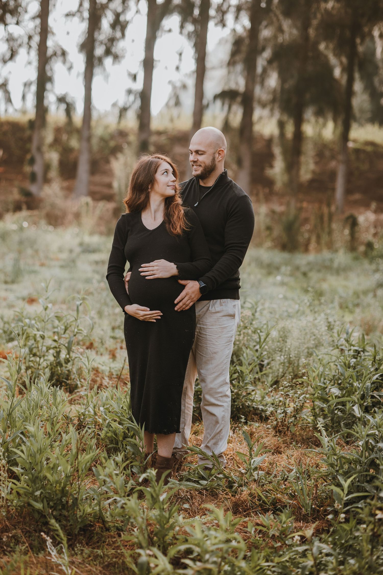 A couple embracing during a maternity photoshoot in a wooded setting at sunset.