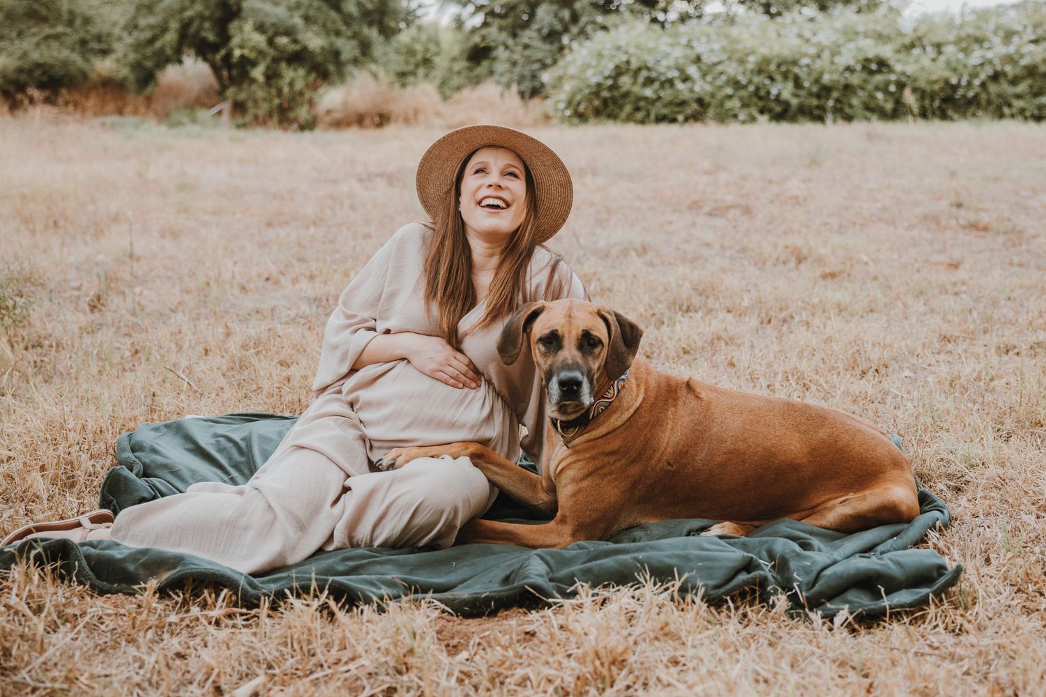 Woman in a hat sits on a blanket with her loyal brown dog in a sunny field.