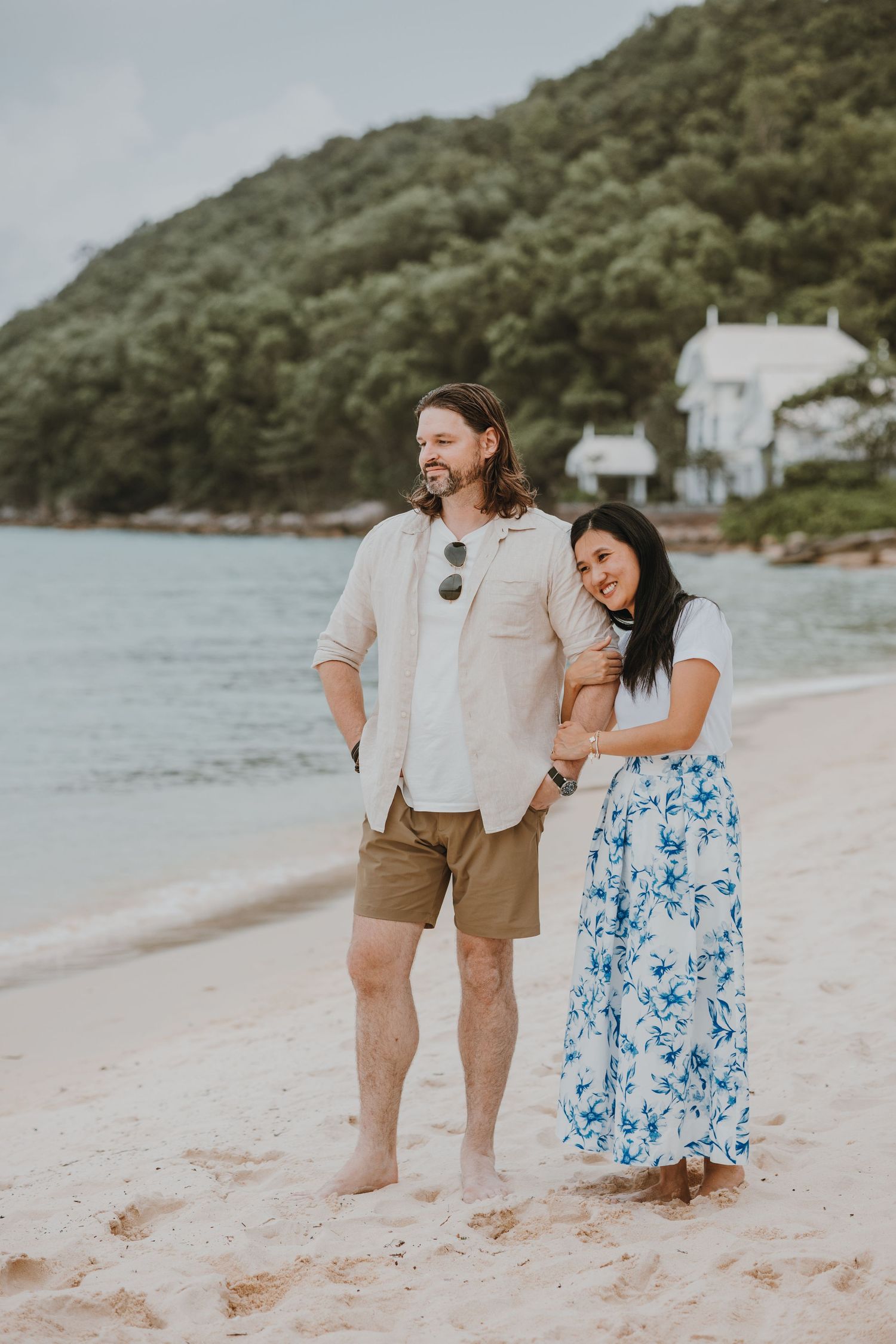 A couple stands together on a sandy beach with green hillside and white houses in the background.