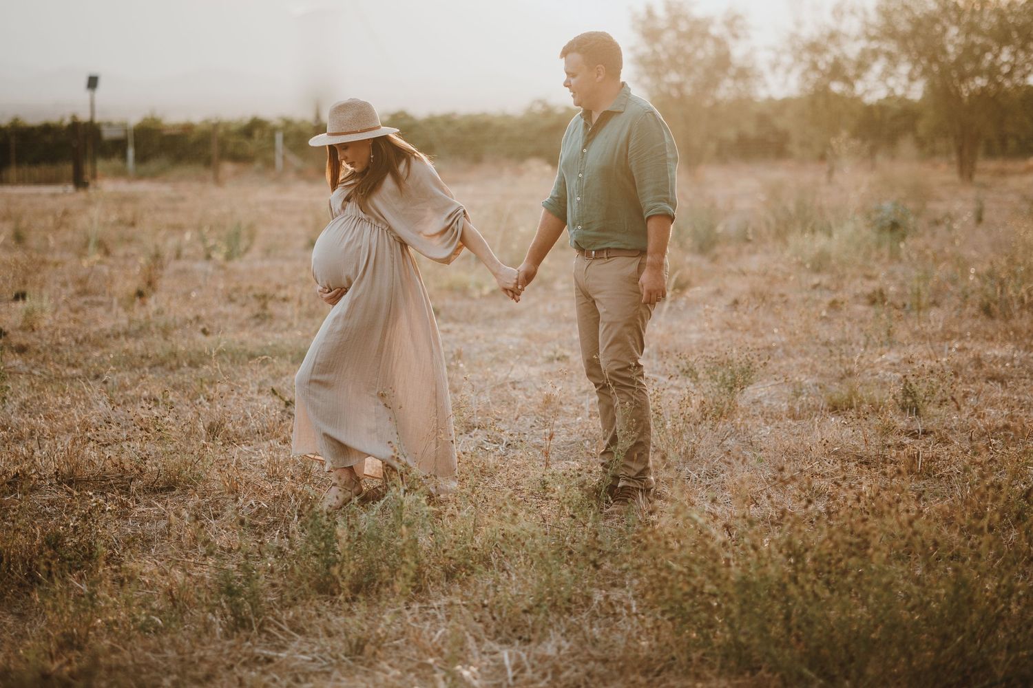 A couple holds hands in a sunlit grassy meadow during golden hour creating a dreamy romantic atmosphere.