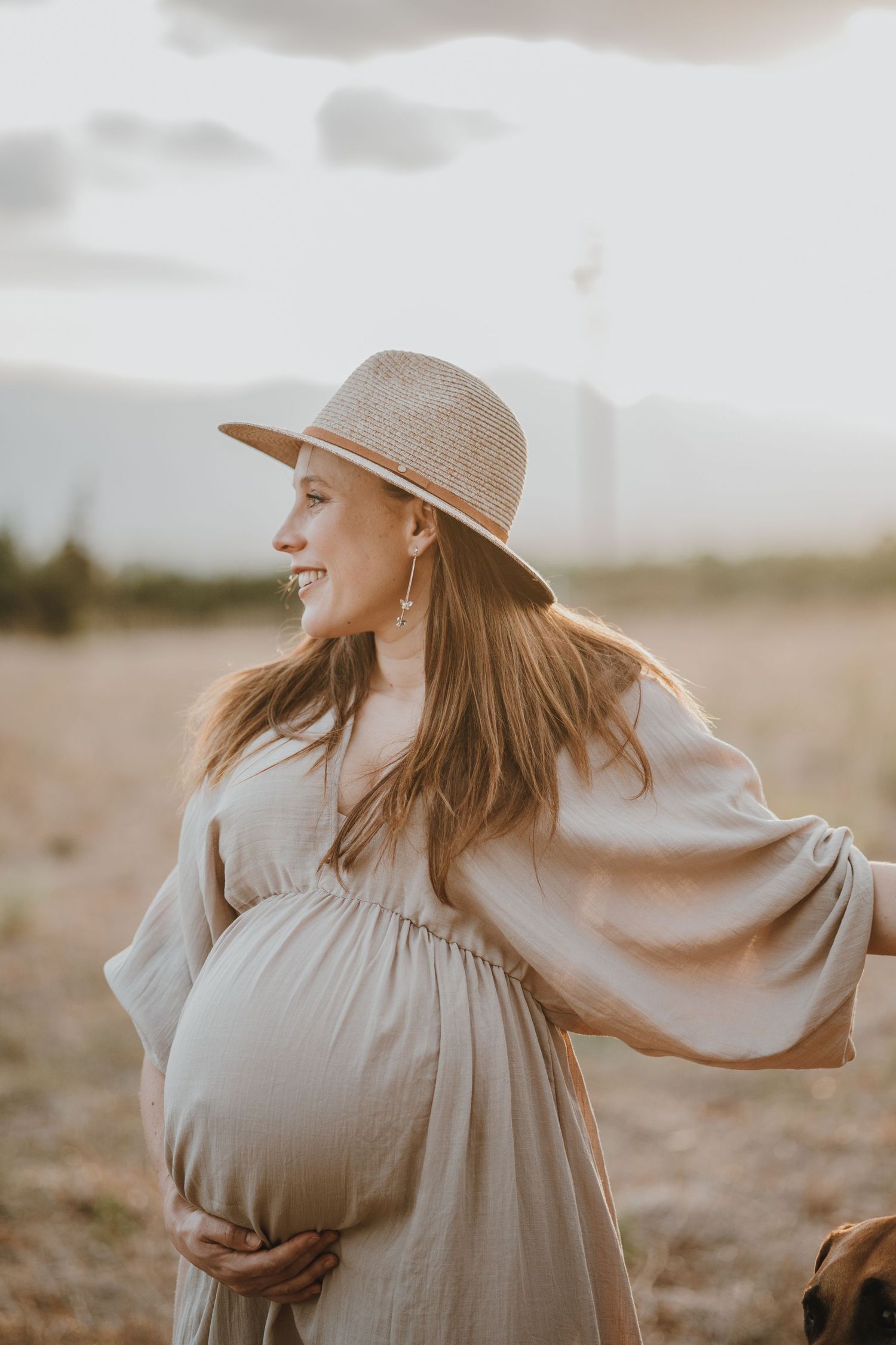 Expectant mother in a flowing dress and hat poses in golden sunlight.