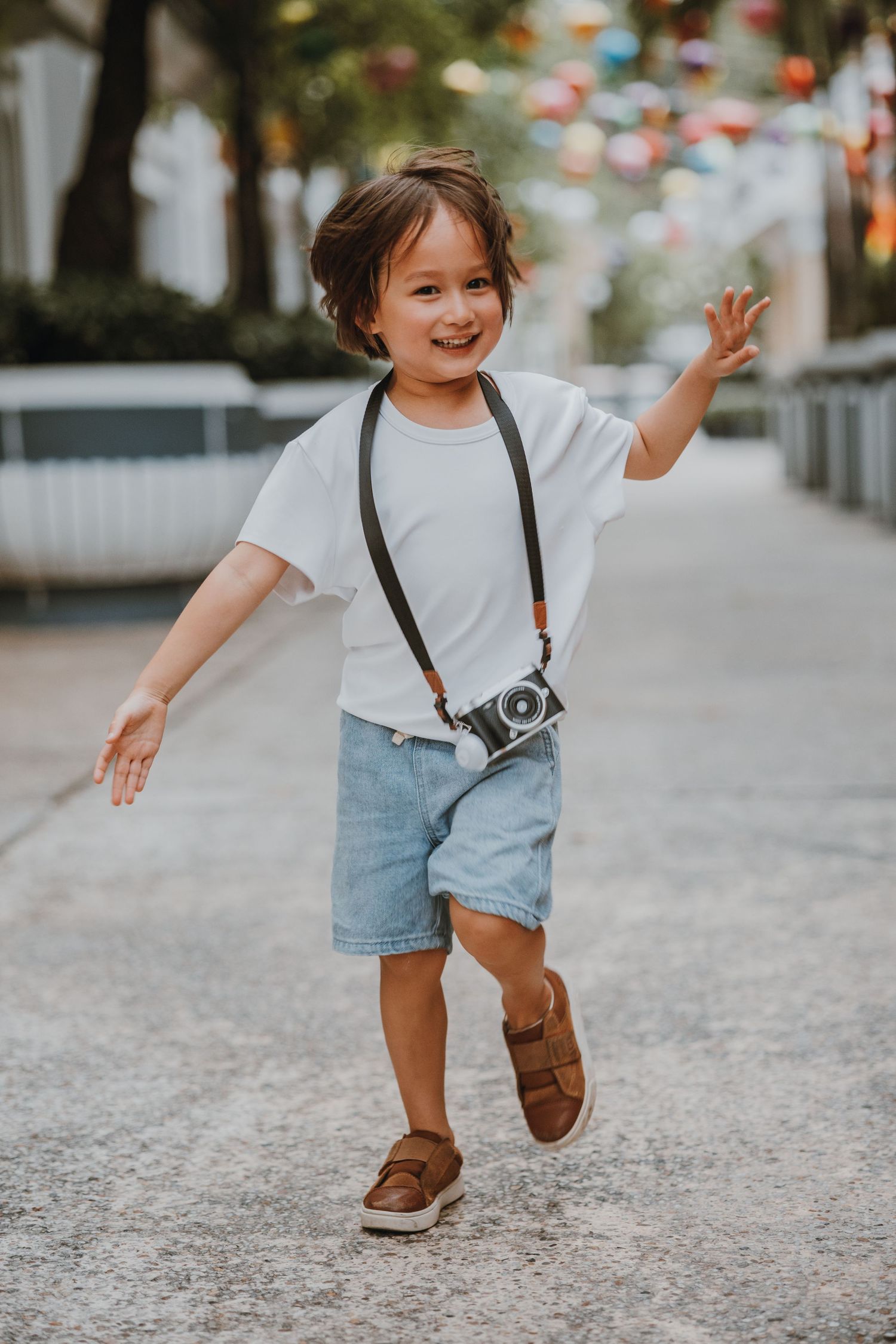Joyful young child in casual outfit with vintage camera running and playing on a path with colorful decorations overhead.
