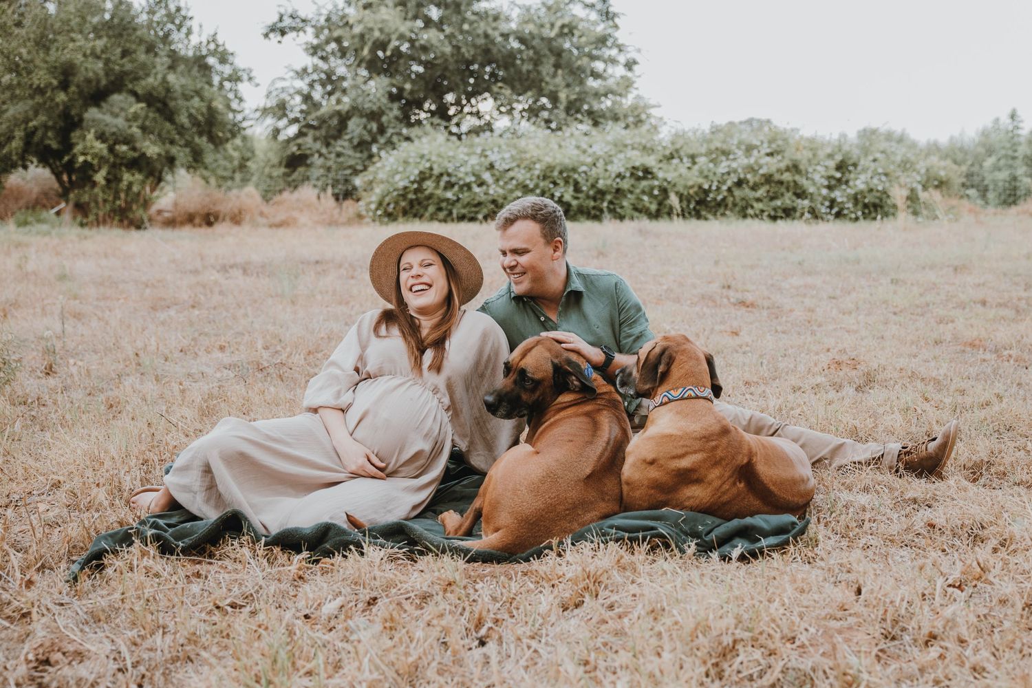 A couple relaxes on a blanket outdoors with their brown dog in a natural setting.