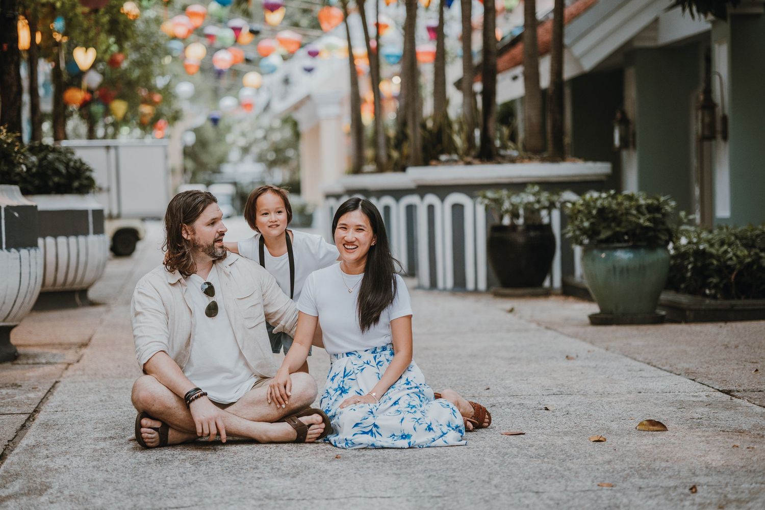 Three young friends sitting together and laughing on an urban sidewalk with potted plants and trees in the background.