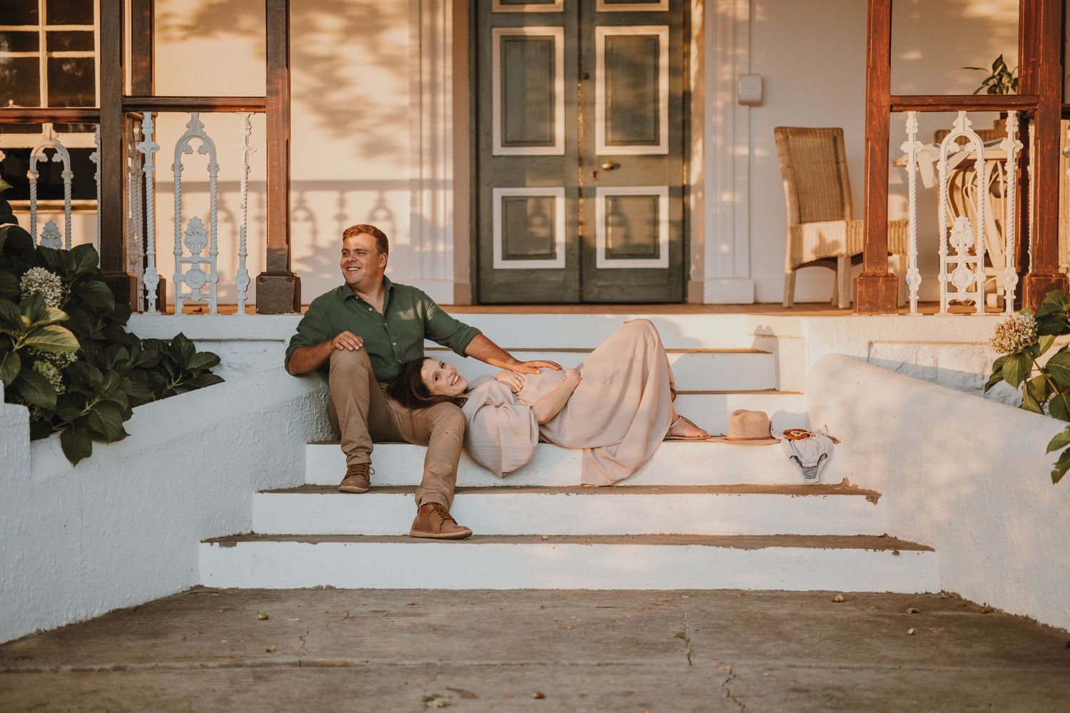 Two people relax on white porch steps during golden hour with warm sunlight casting shadows on a Mediterranean-style facade.