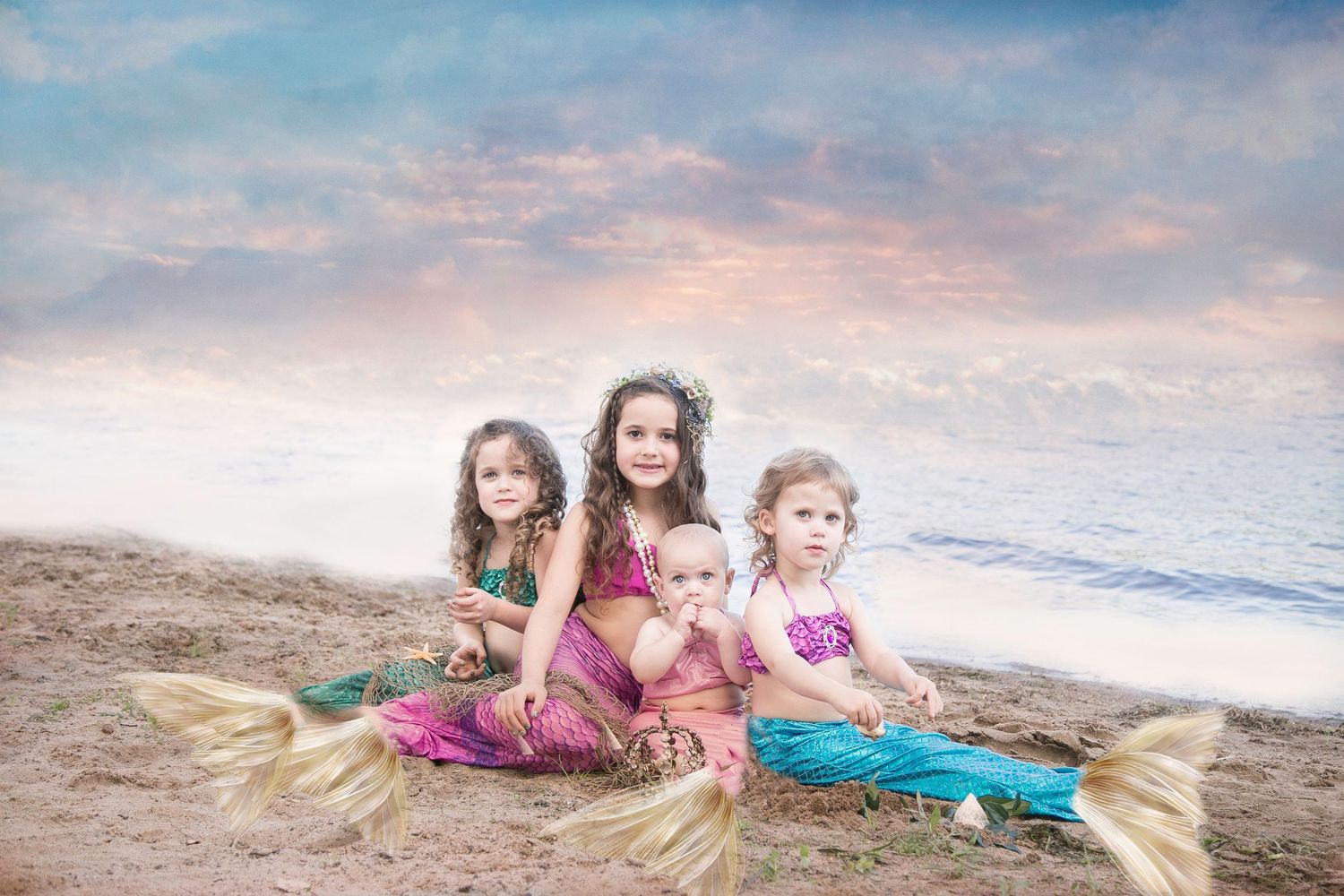 Four young girls dressed as mermaids posing on a beach with a colorful sunset sky background.