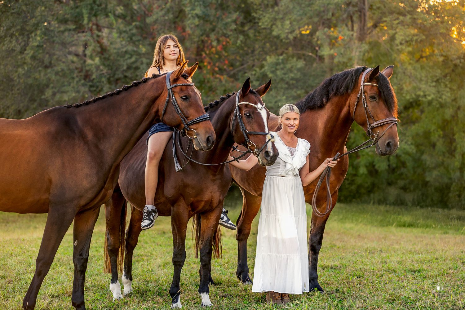 Two beautiful brown horses stand in a grassy field at sunset while riders in white dresses pose with them.