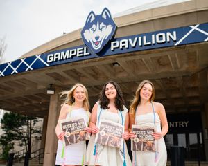 Three graduates pose with UConn memorabilia outside the Gampel Pavilion entrance featuring the Husky mascot logo.
