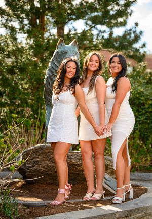 Three graduates in white dresses pose by a Husky statue on UConn campus during golden hour.